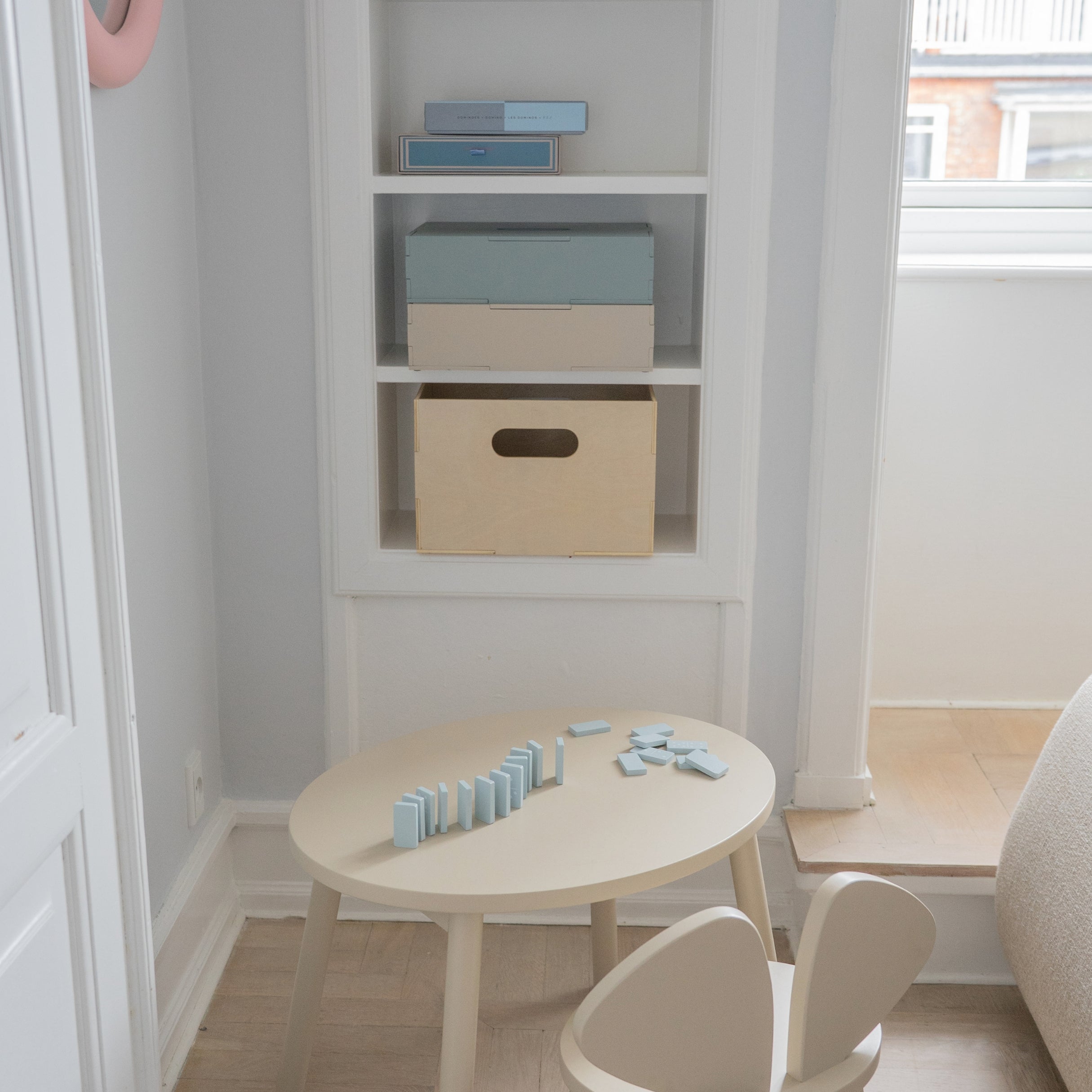 Children's play area with a round table and chairs, shelves with boxes, and a window in the background.