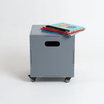 A gray metal rolling storage cabinet with casters and a handle cutout, shown on a white studio background with a stack of colorful books on top.