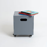 A gray metal rolling storage cabinet with casters and a handle cutout, shown on a white studio background with a stack of colorful books on top.