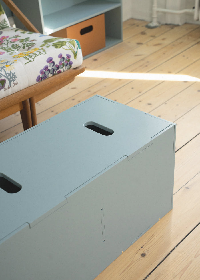 Olive green Nofred Cube Long storage bench with its lid fitted, viewed from above on a wooden floor beside a floral-upholstered chair.