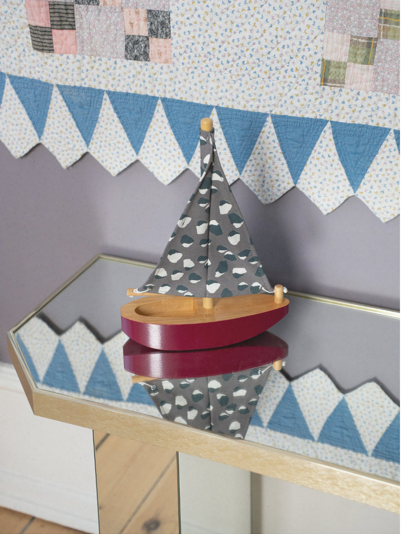 Nofred Harbour Boat in burgundy with a patterned grey sail, displayed on a mirrored table beneath a patchwork wall hanging.