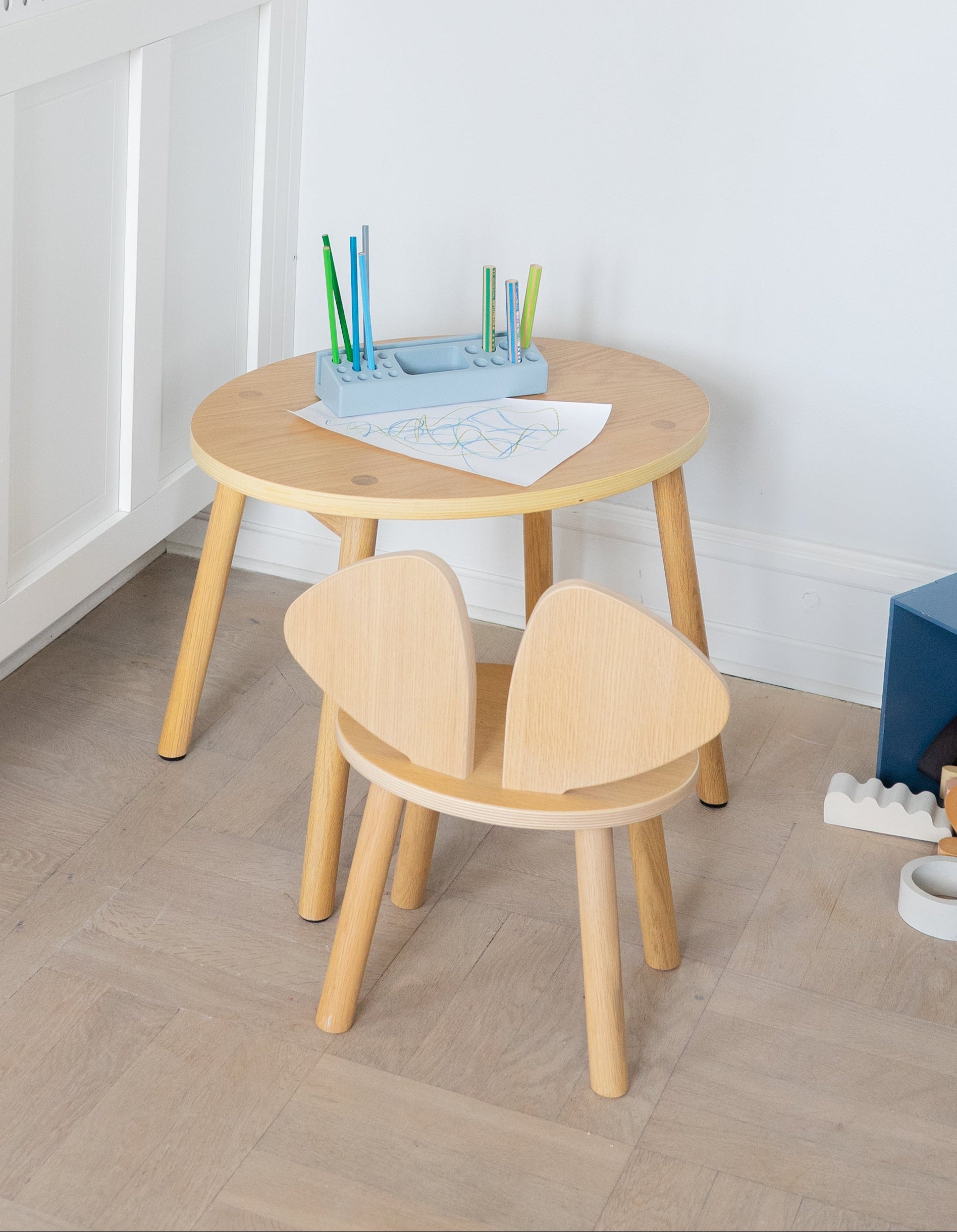 A small children's play area in a bright room, featuring a round light wood table, two matching wooden chairs, a blue storage box, and a white wall-mounted cabinet.
