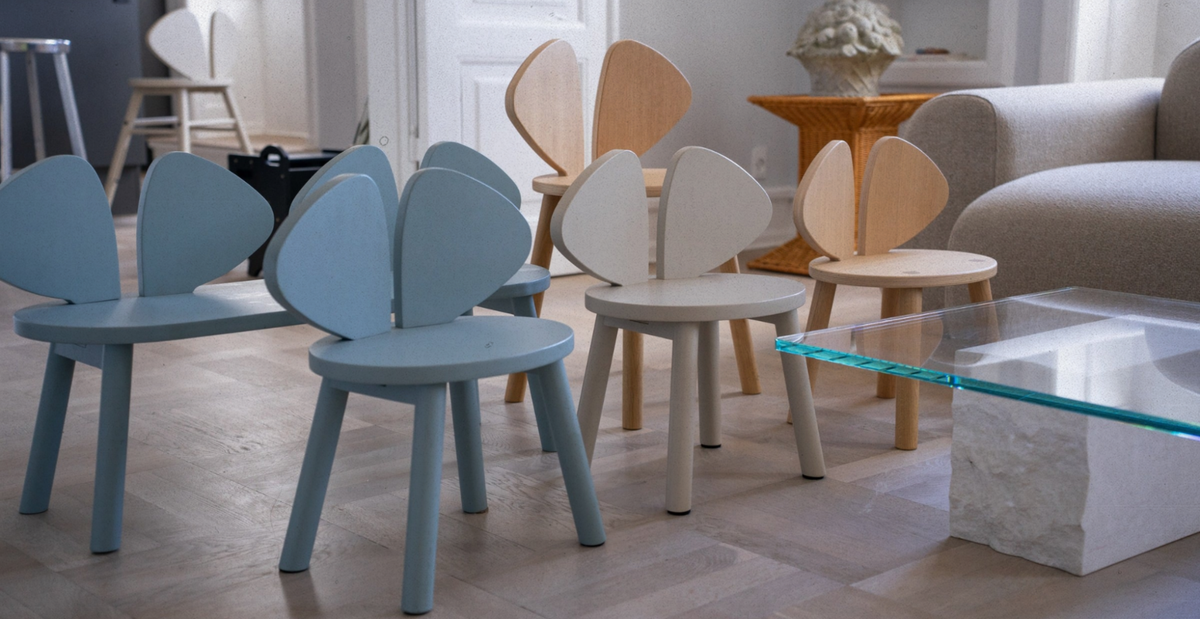 A group of modern wooden chairs with heart-shaped backs in light blue, cream, and natural wood tones, arranged in a bright living room next to a glass-top coffee table.