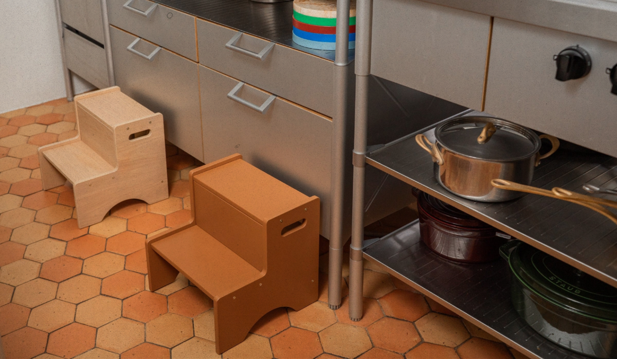 Two wooden two-step stools in natural and medium brown tones placed on a terracotta hexagonal tile floor next to stainless steel kitchen cabinets and a shelf with pots.