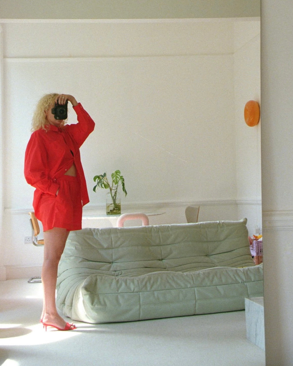 Person in a red outfit taking a mirror selfie in a minimalistic room with a green sofa.