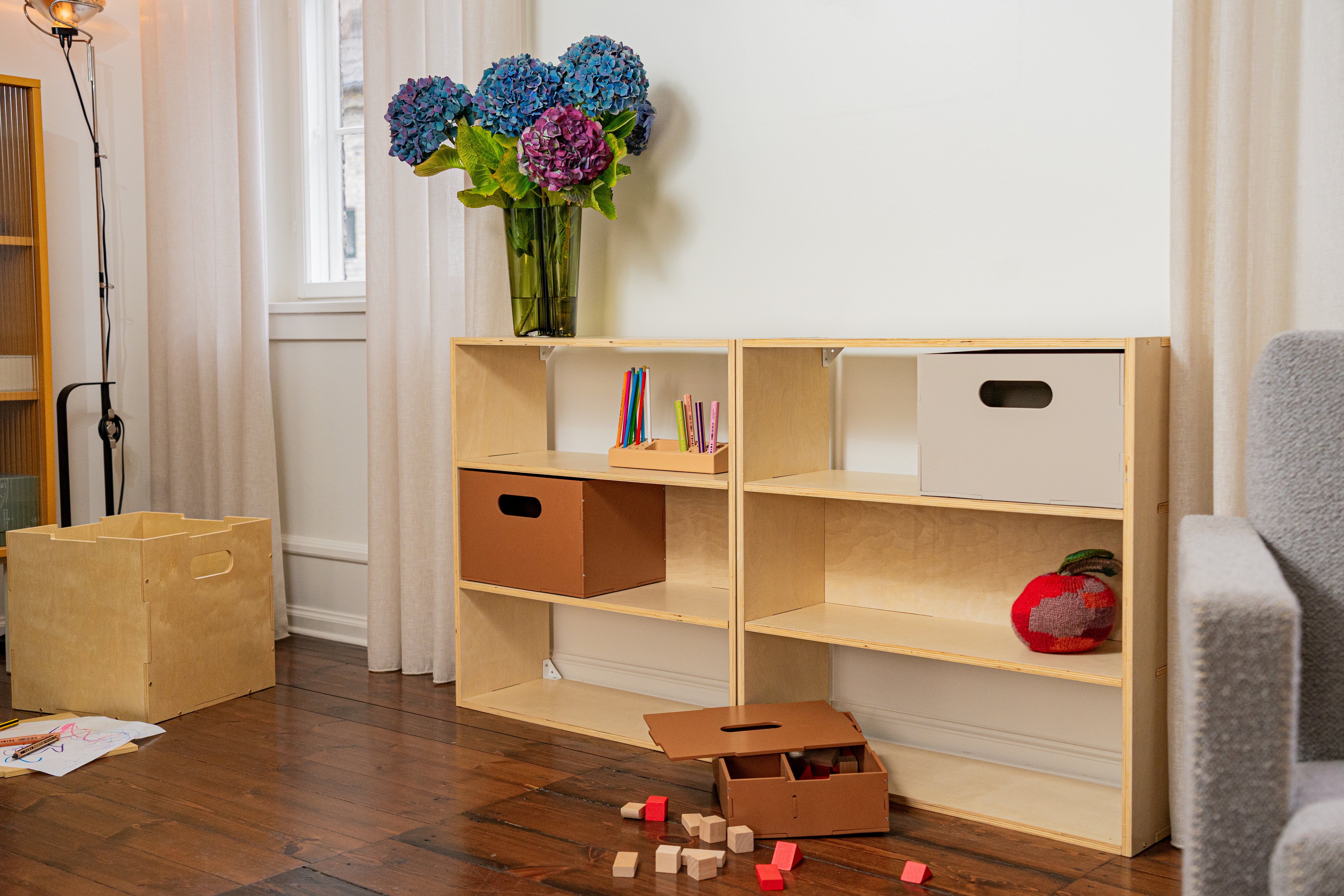 A light natural wood modular children's bookshelf with open shelves and storage cutouts, styled in a child's room with a vase of blue flowers, cardboard bins, and blocks on a wooden floor.
