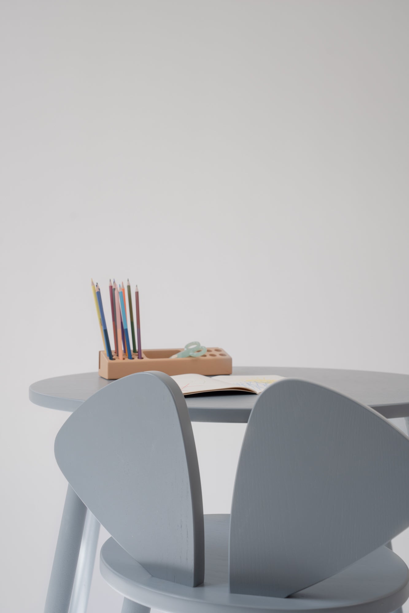 A minimalist light gray children's table and chair set with a small wooden pencil holder on the table, set against a plain light gray wall.