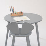 A minimalist children's table set featuring a round gray table and two matching gray chairs, styled with a small wooden pencil holder and an open book on a clean white background.