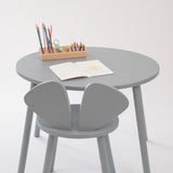 A minimalist children's table set featuring a round gray table and two matching gray chairs, styled with a small wooden pencil holder and an open book on a clean white background.