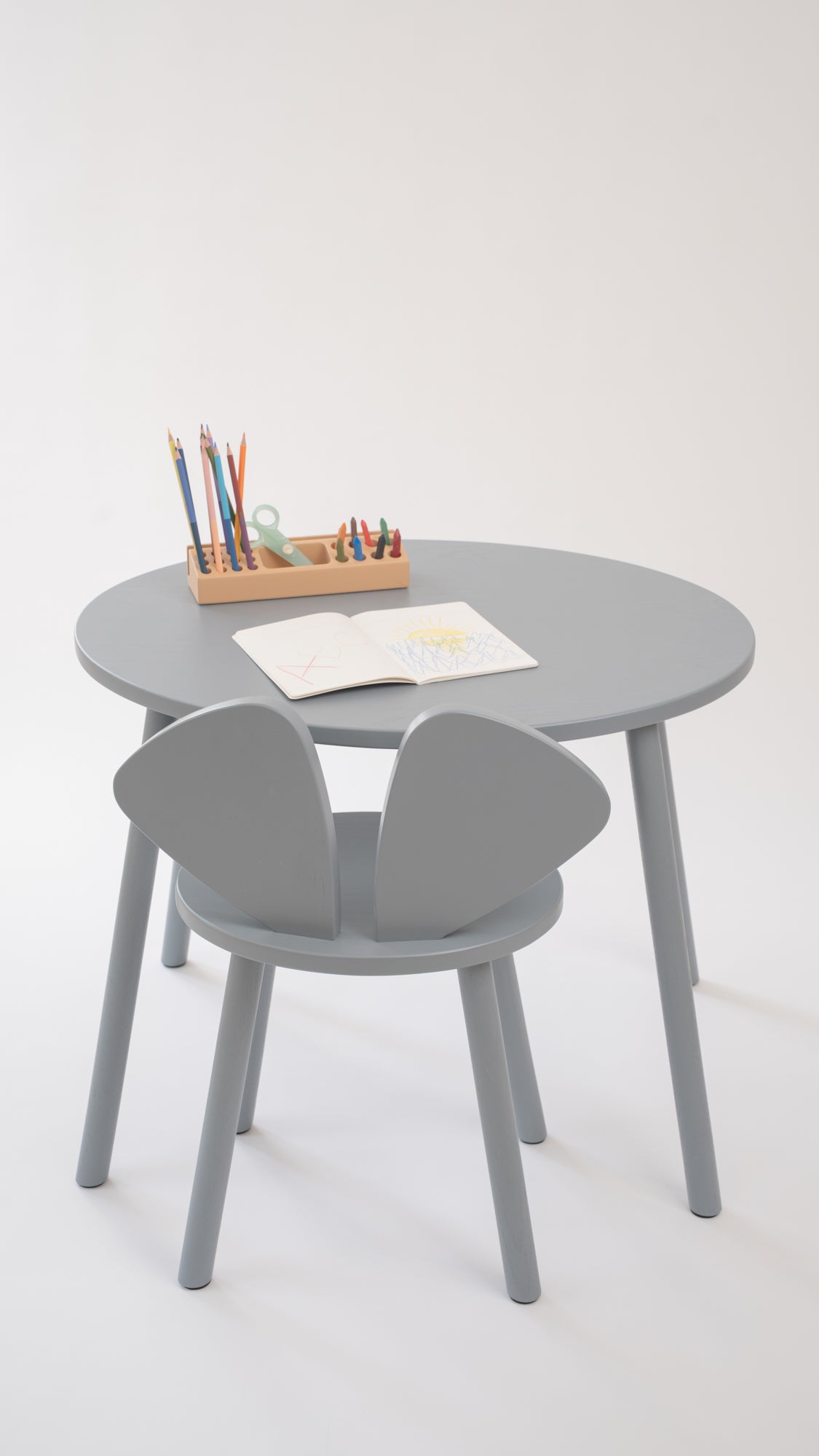 A minimalist children's table set featuring a round gray table and two matching gray chairs, styled with a small wooden pencil holder and an open book on a clean white background.