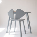A light gray wooden dining chair with a unique wing-shaped backrest, shown next to a matching light gray wooden table in a minimalist studio setting.