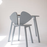 A light gray wooden dining chair with a unique wing-shaped backrest, shown next to a matching light gray wooden table in a minimalist studio setting.