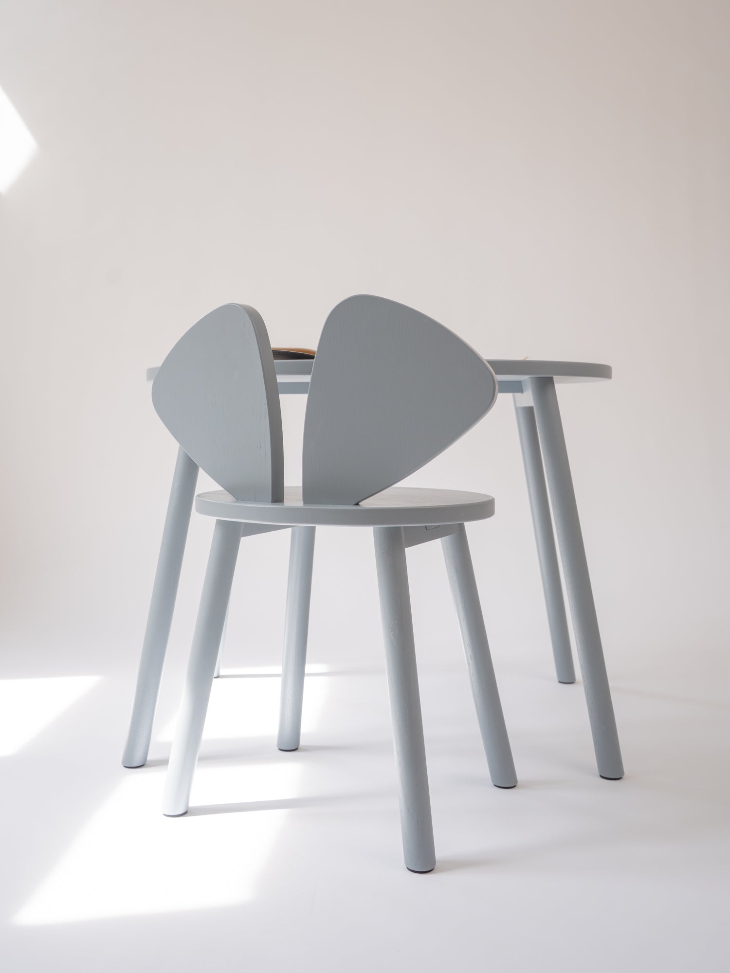 A light gray wooden dining chair with a unique wing-shaped backrest, shown next to a matching light gray wooden table in a minimalist studio setting.