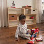 Child playing with toy fire trucks on a wooden floor in a room with a bookshelf.