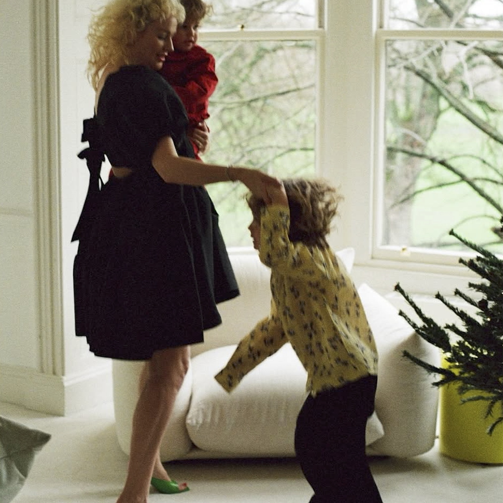 Woman in a black dress interacting with two children in a bright room with large windows.