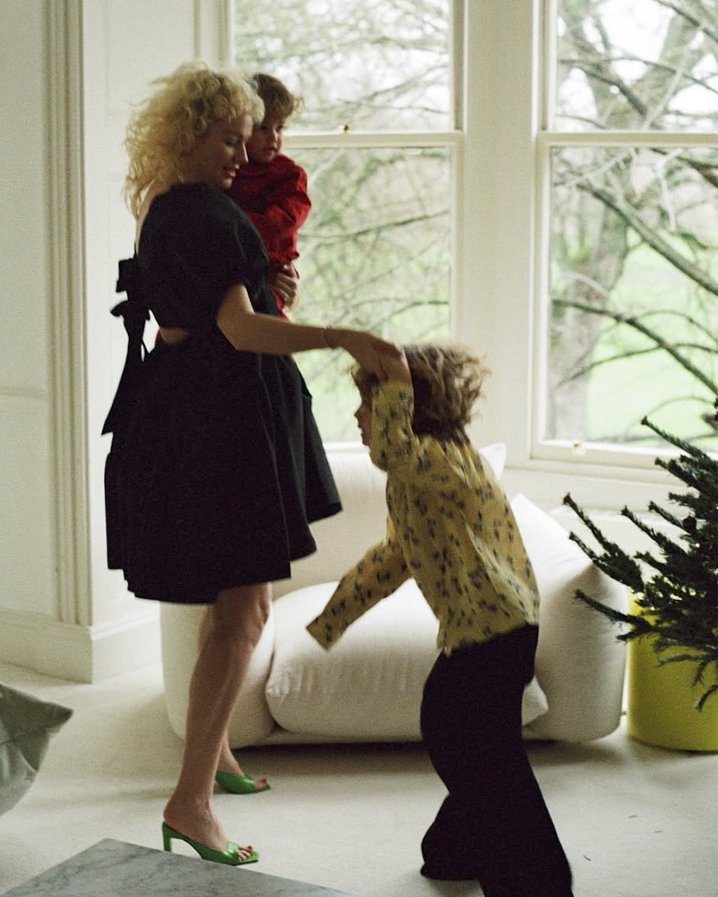 Woman in a black dress interacting with two children in a bright room with large windows.