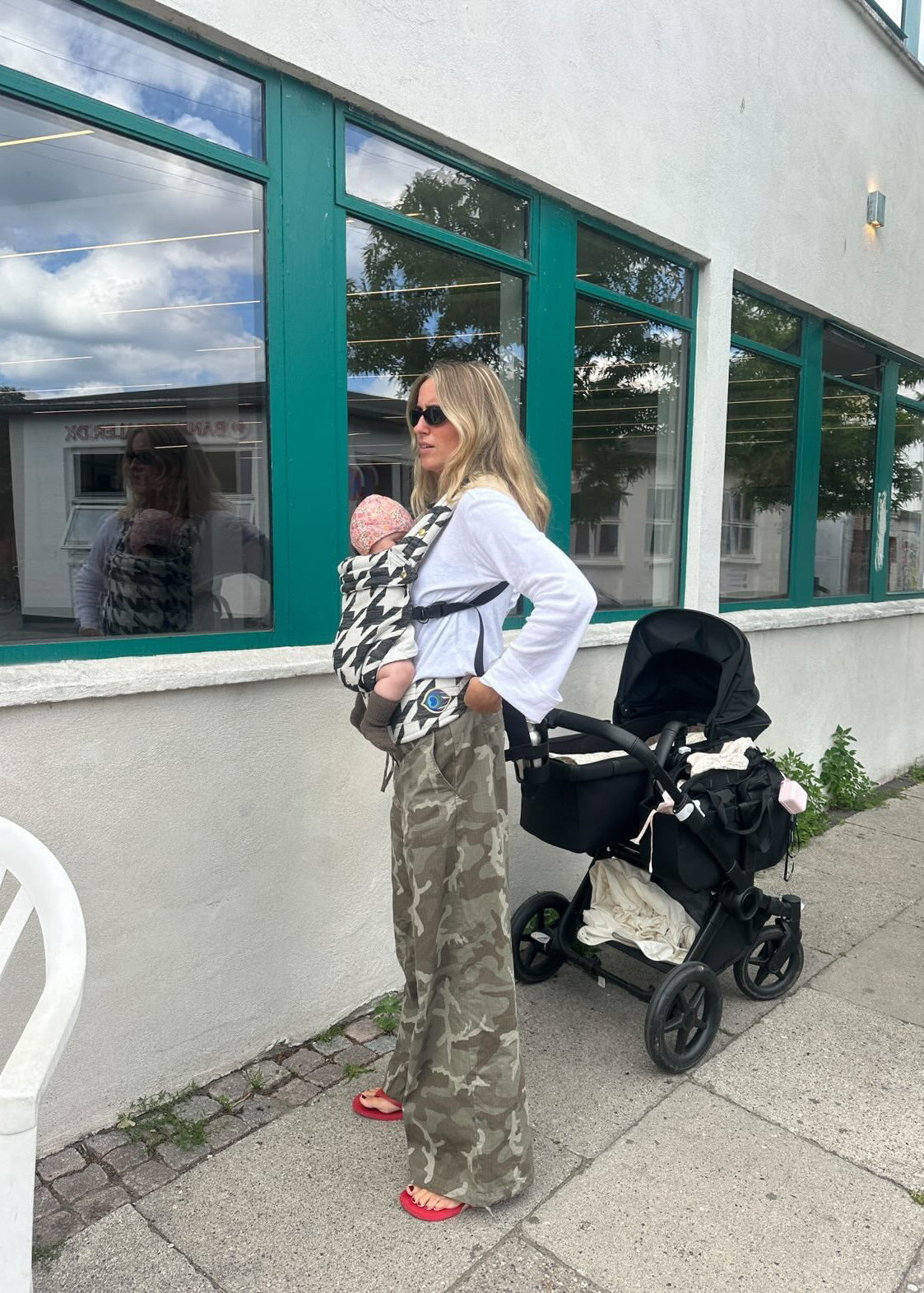A woman wearing a black and white patterned baby carrier with a baby inside, standing outdoors next to a black stroller. She is wearing a white long-sleeve top, camouflage pants, and red sandals in front of a building with green-framed windows.