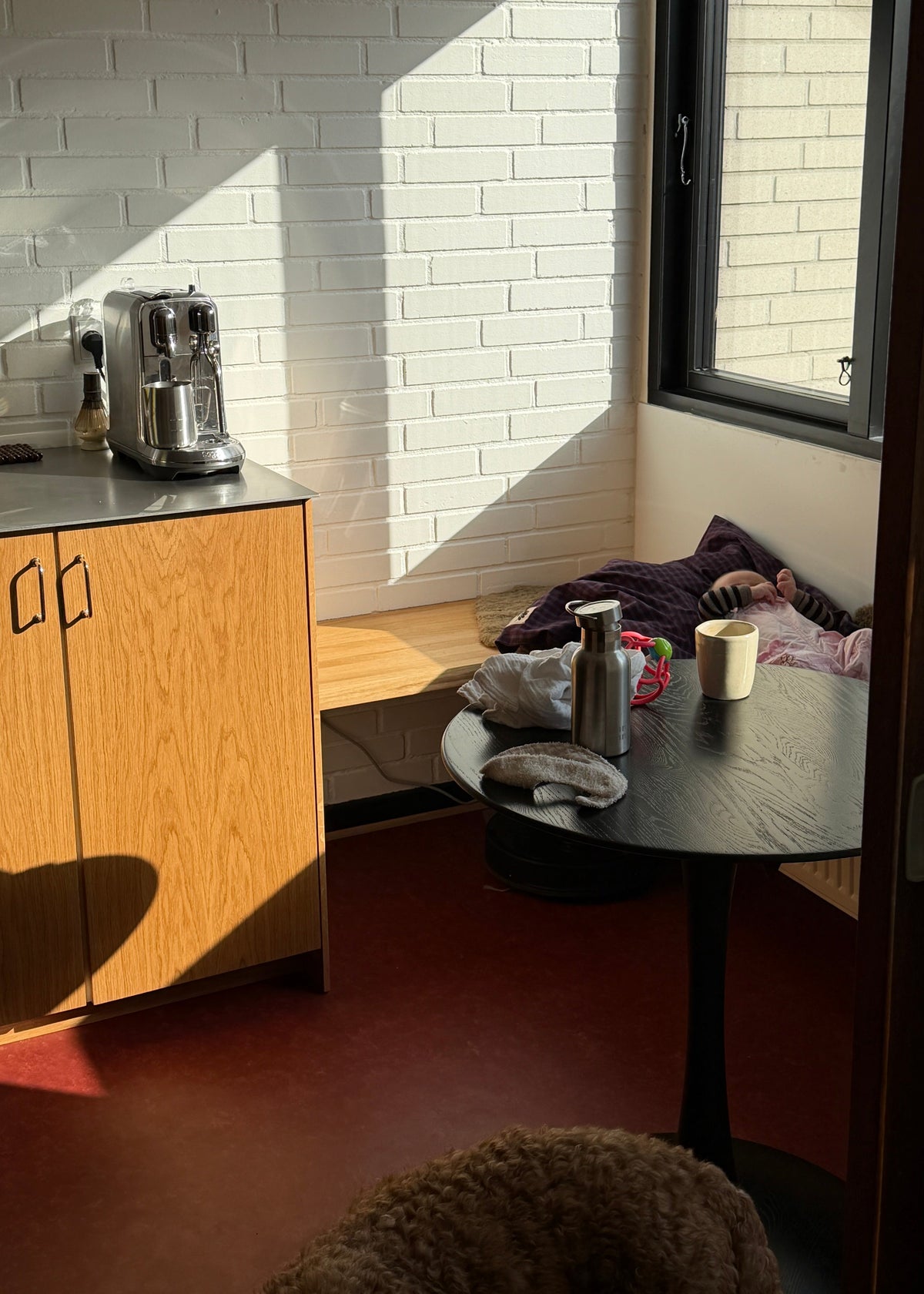 A cozy interior corner with a white brick wall, warm sunlight, a small wooden cabinet, a round black table with a metal bottle and mug, and a dog partially visible in the foreground.
