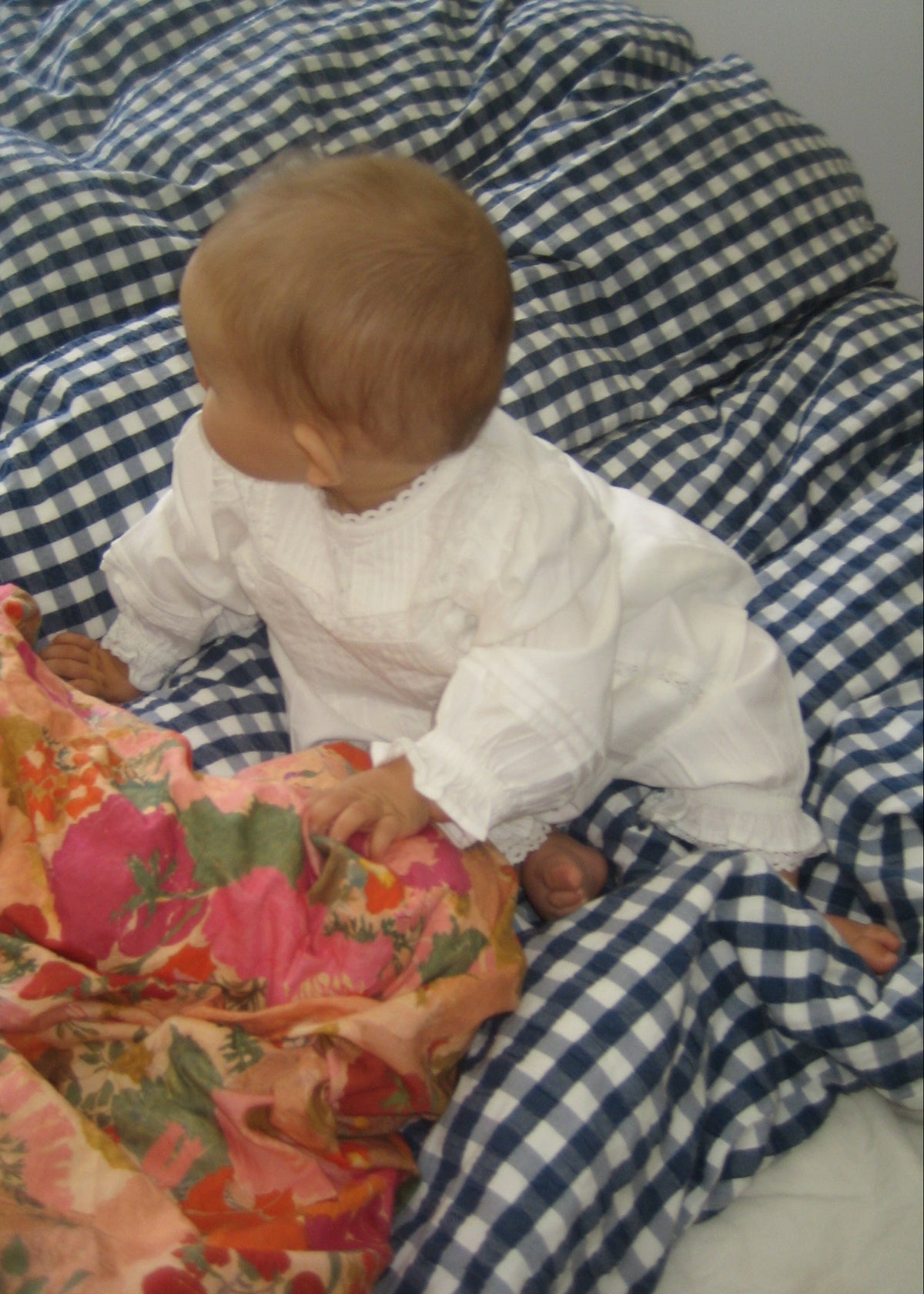 A baby wearing a white long-sleeve smocked romper sits on a blue and white gingham blanket, holding a pink and orange floral patterned fabric.