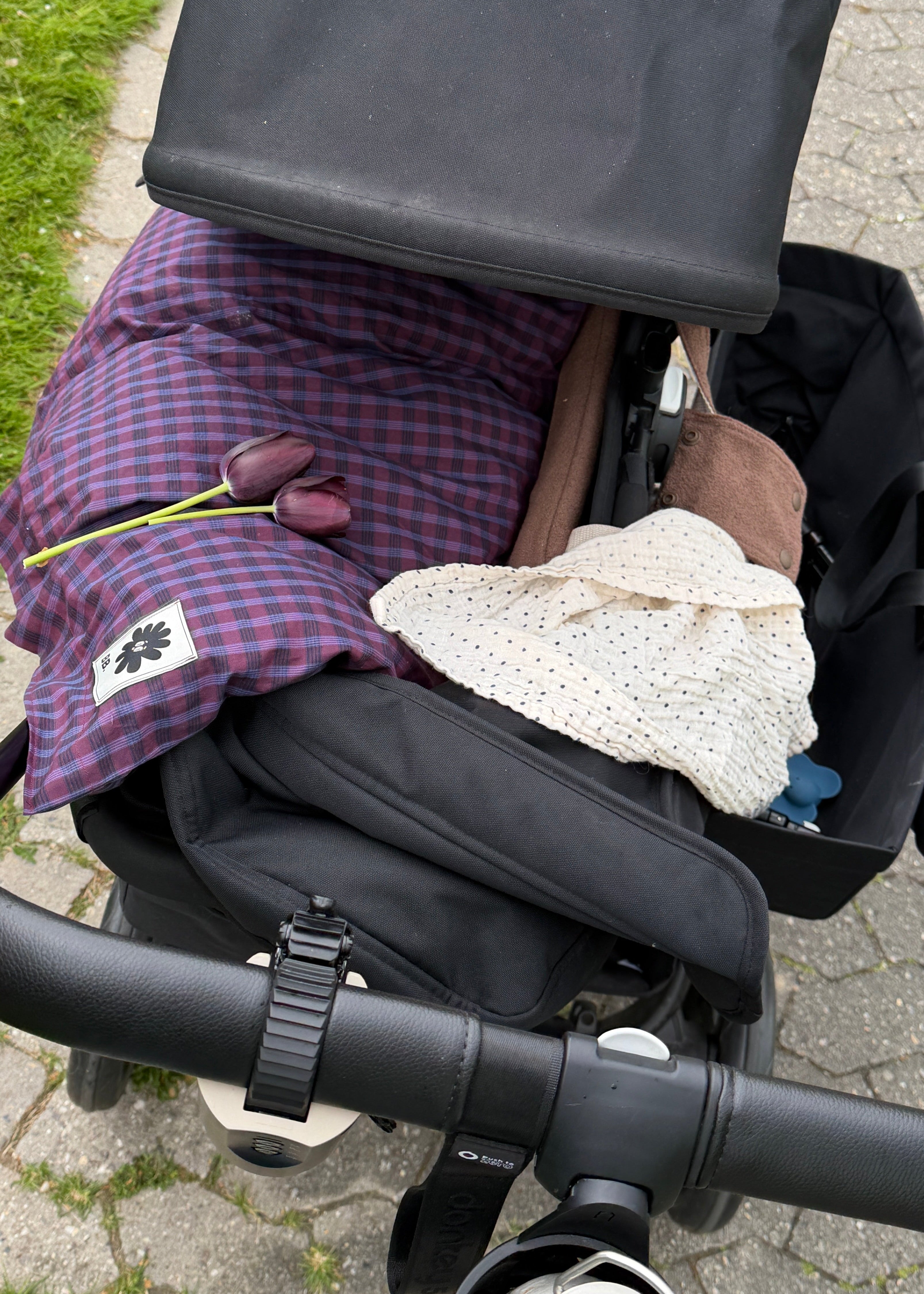 A close-up view of a black stroller or baby carrier outdoors on a paved path, with a purple plaid blanket, a cream knit hat, and other baby items packed inside.