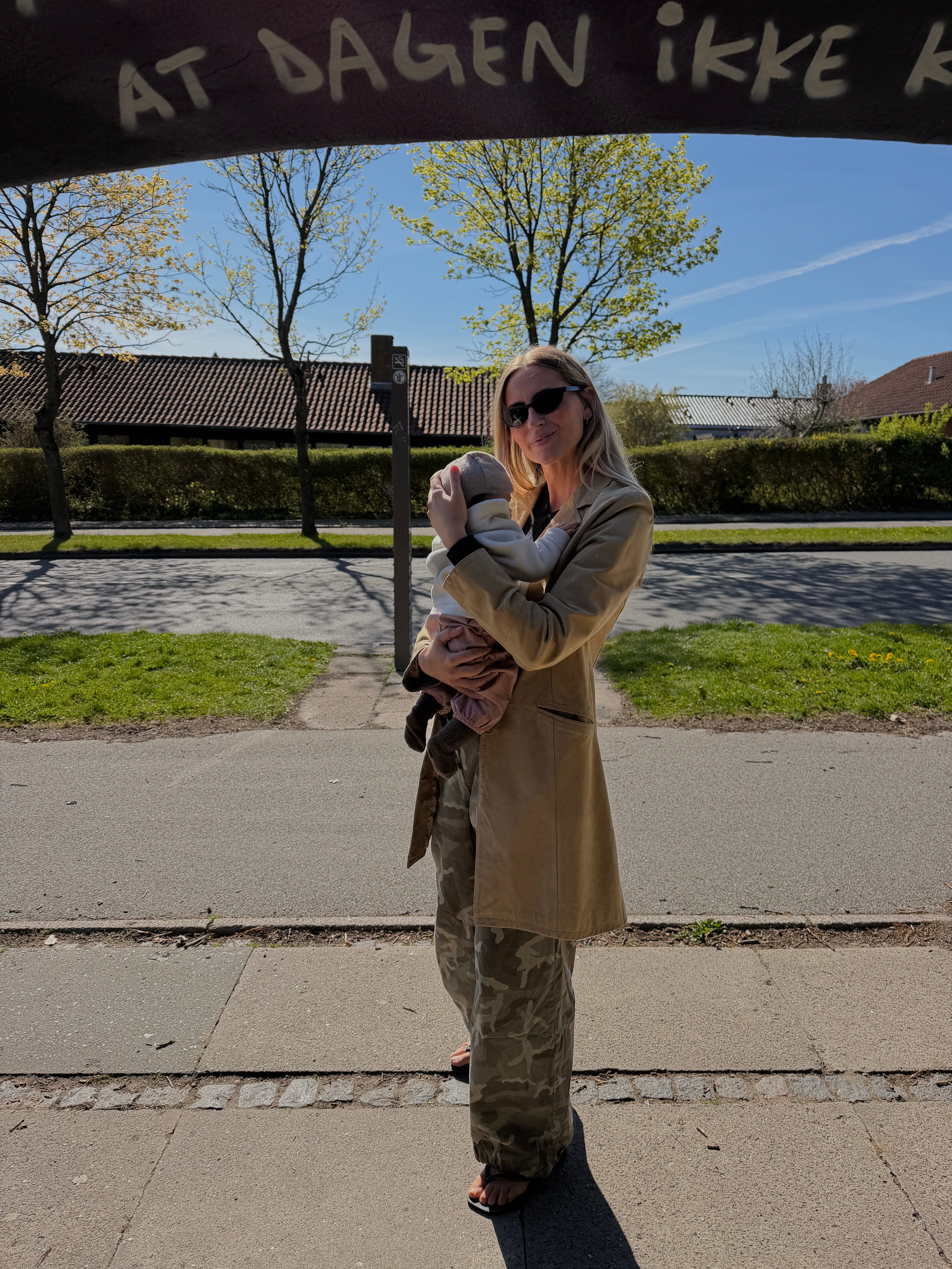 A woman wearing a beige trench coat and camouflage pants holds a baby outdoors under a dark sign that reads 'AT DAGEN IKKE R'.