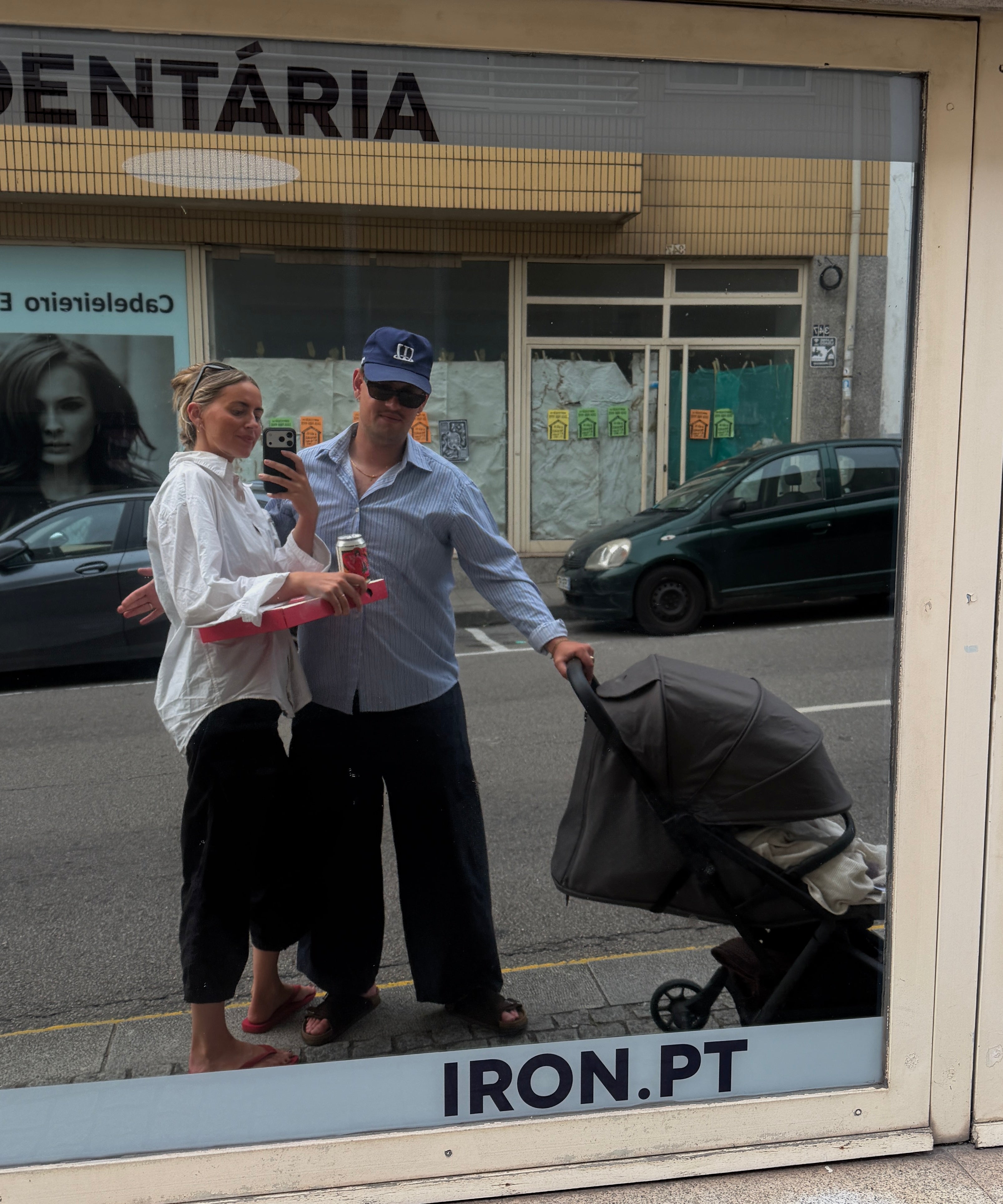A couple standing outside a storefront window next to a stroller, reflected in the glass. The storefront has the text 'DENTÁRIA' and 'IRON.