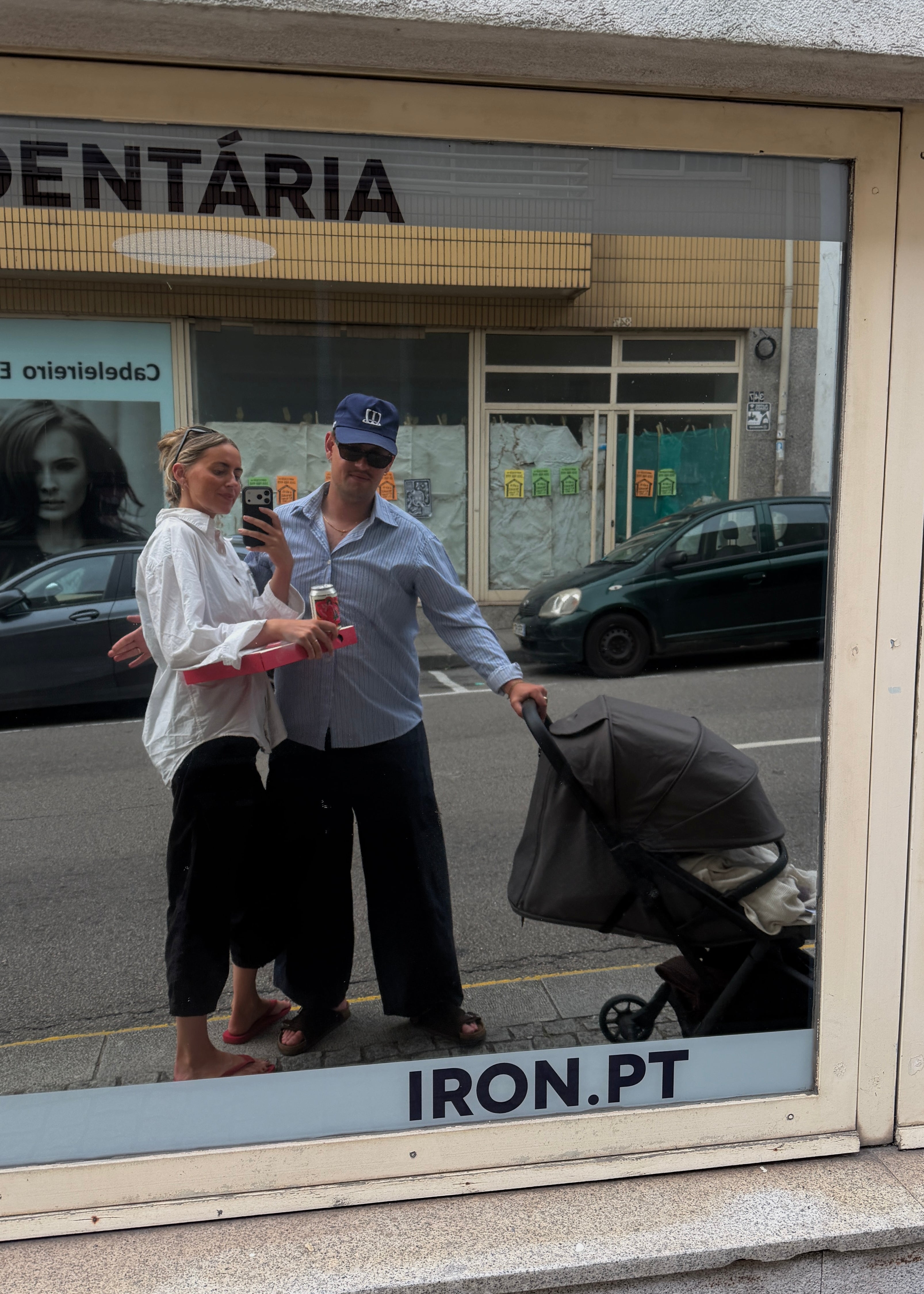 A couple standing outside a storefront window next to a stroller, reflected in the glass. The storefront has the text 'DENTÁRIA' and 'IRON.