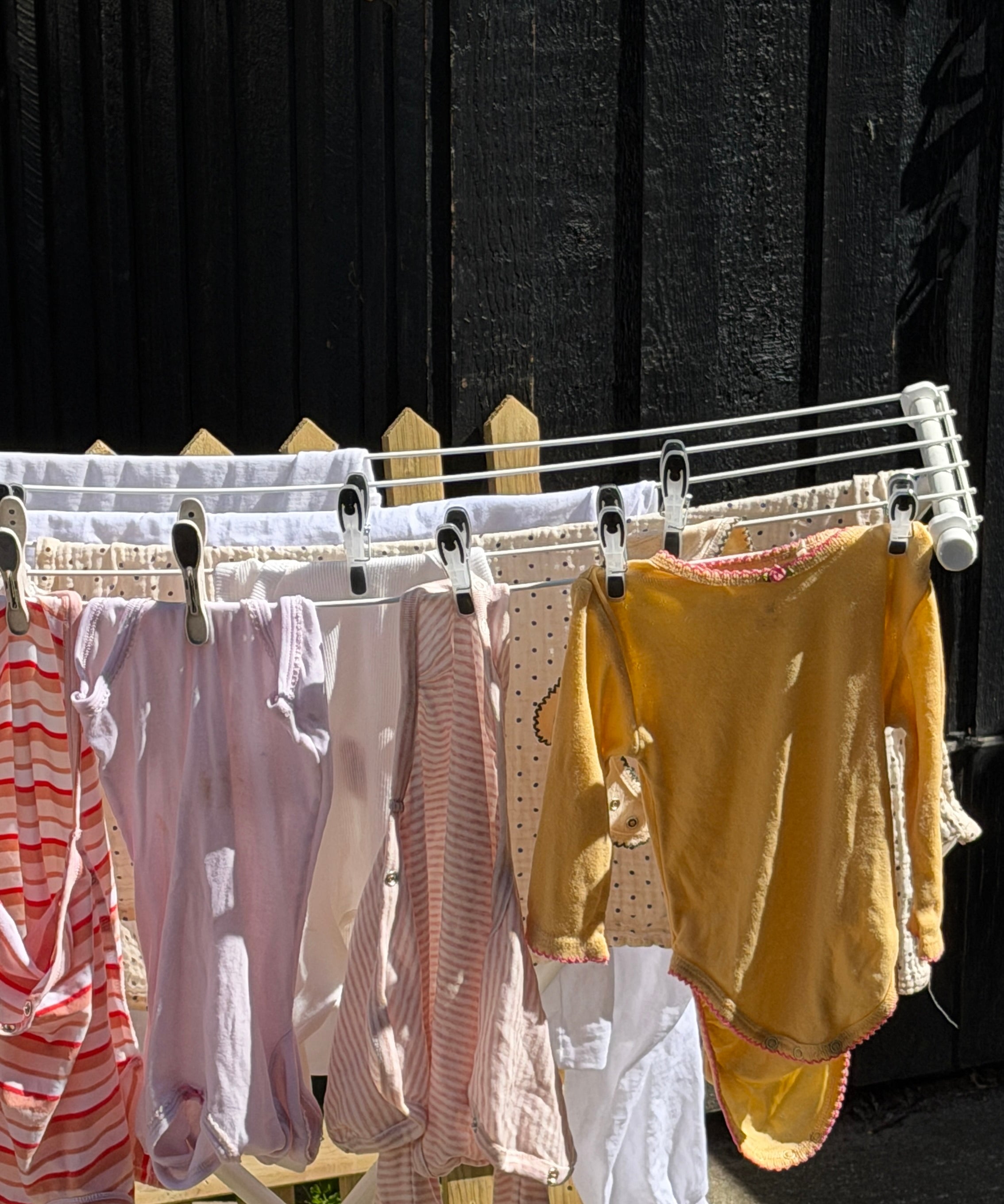 A white folding clothesline draped outdoors against a dark wooden fence, holding several baby garments including striped pink and white tops, a pale pink dress, a mustard yellow long-sleeve top, and white fabric pieces.