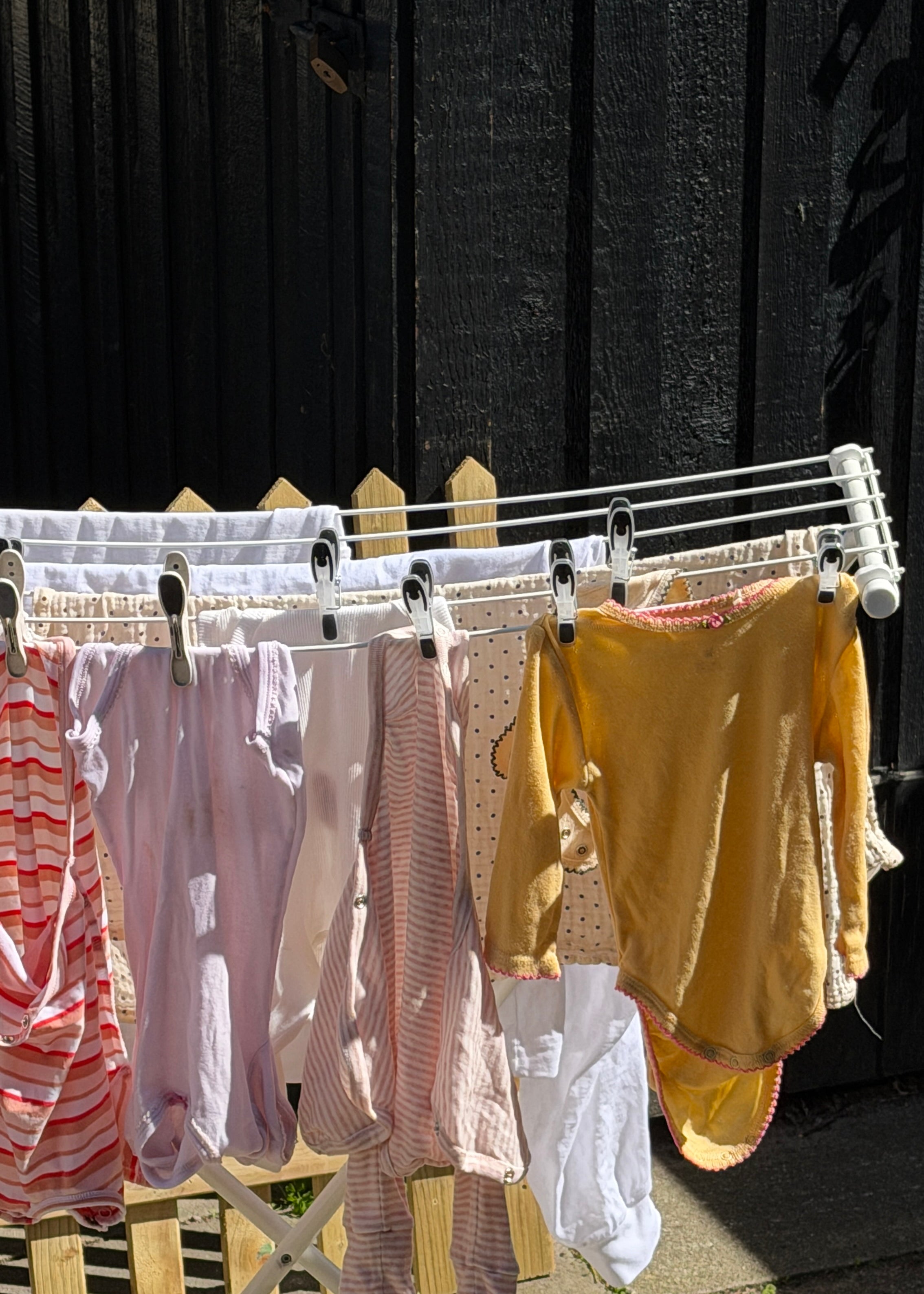 A white folding clothesline draped outdoors against a dark wooden fence, holding several baby garments including striped pink and white tops, a pale pink dress, a mustard yellow long-sleeve top, and white fabric pieces.