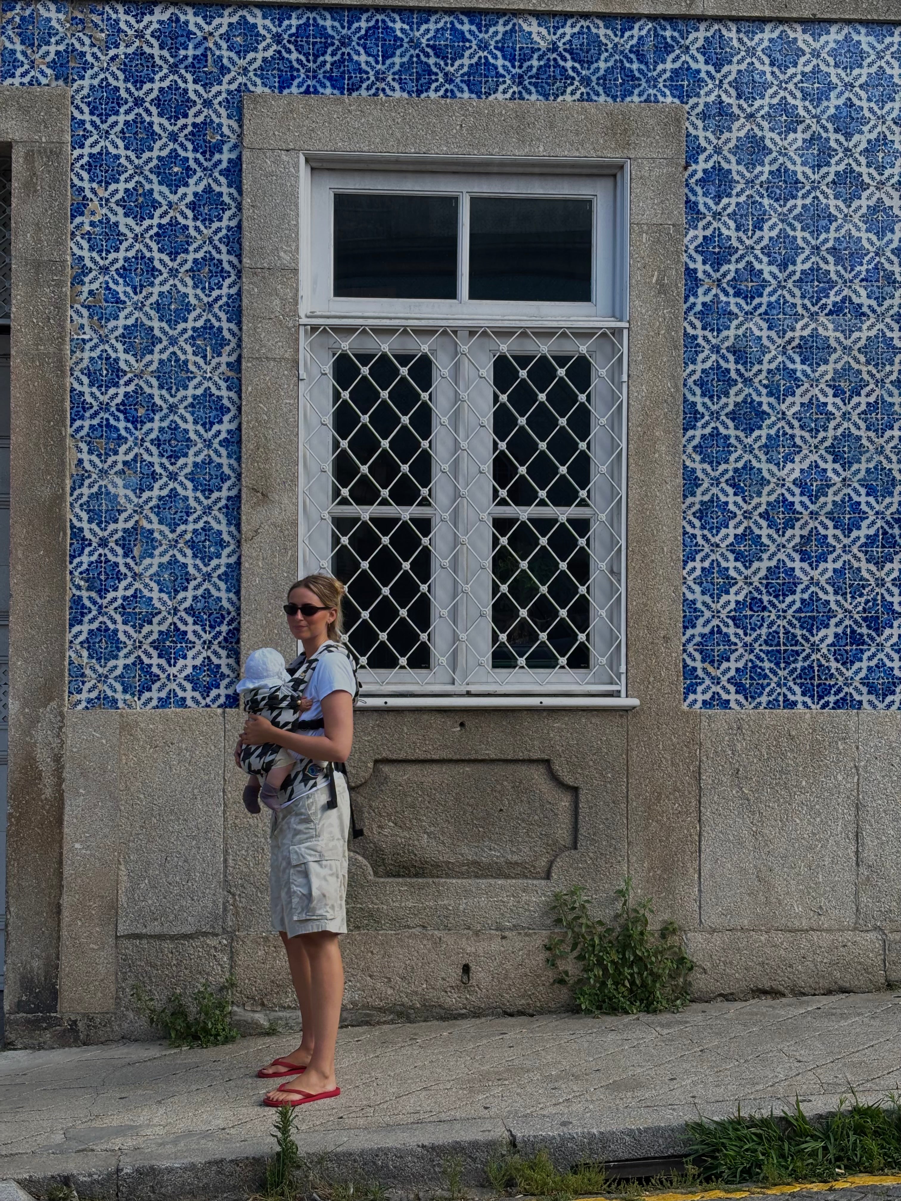 A woman wearing sunglasses, a light blue patterned top, and beige shorts stands on a sidewalk holding a baby carrier with a baby inside, positioned in front of a building with blue and white patterned tiles and a stone window frame.