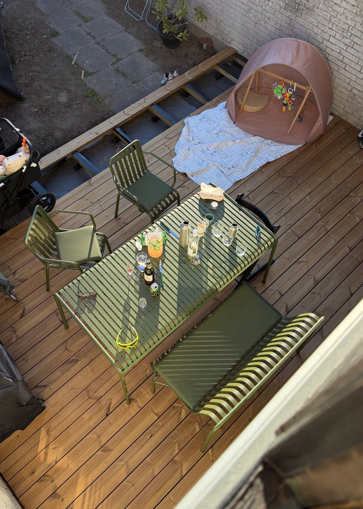 An overhead view of a wooden deck patio set with a green slatted dining table, four green chairs, and a matching green-and-white striped bench. A pink dome play tent is pitched on a white tarp near a white brick wall.