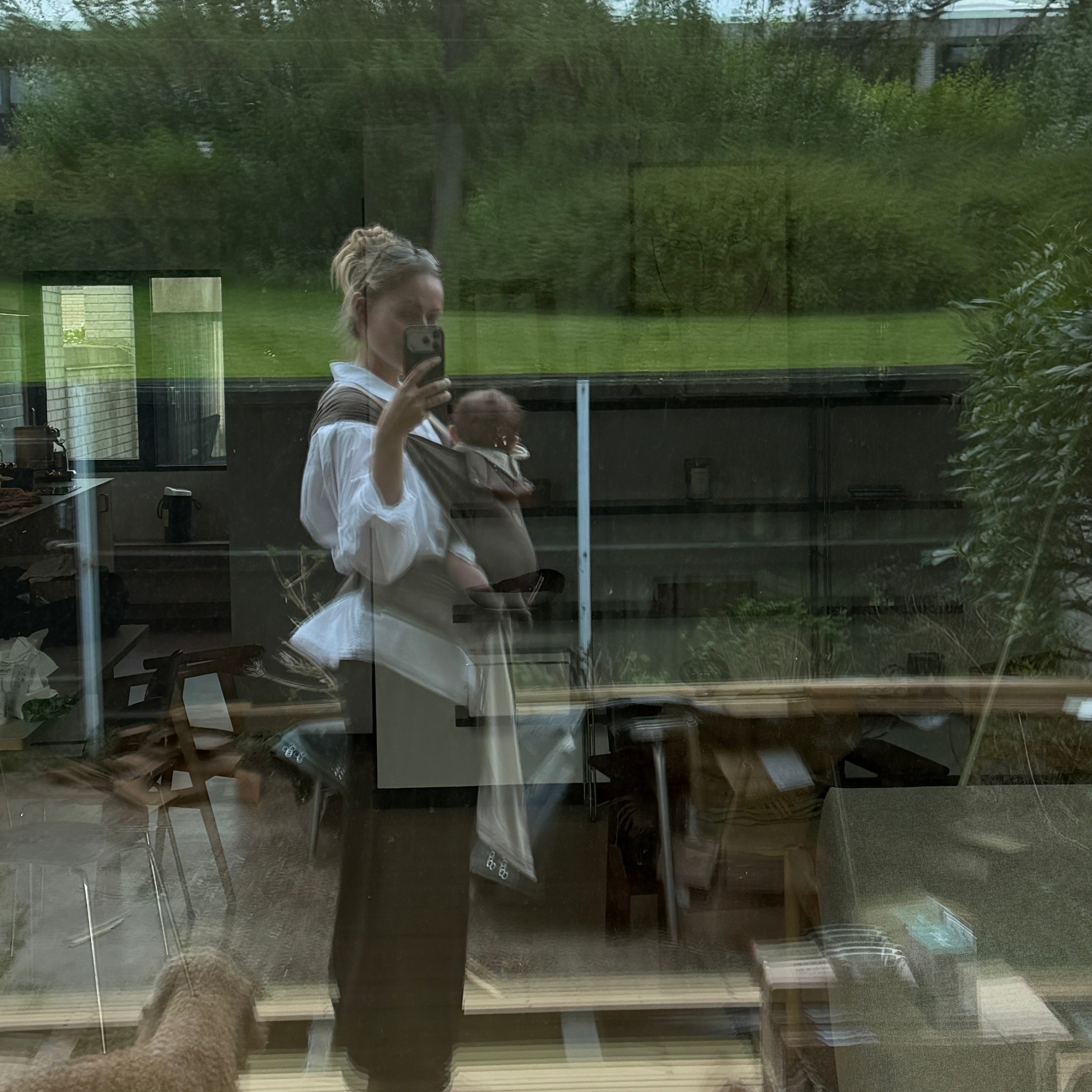 A woman wearing a white blouse and dark skirt stands inside a glass-front room, holding a baby carrier with a baby inside. Large windows behind her show a green outdoor garden, and there are reflections and plastic sheeting on the glass.