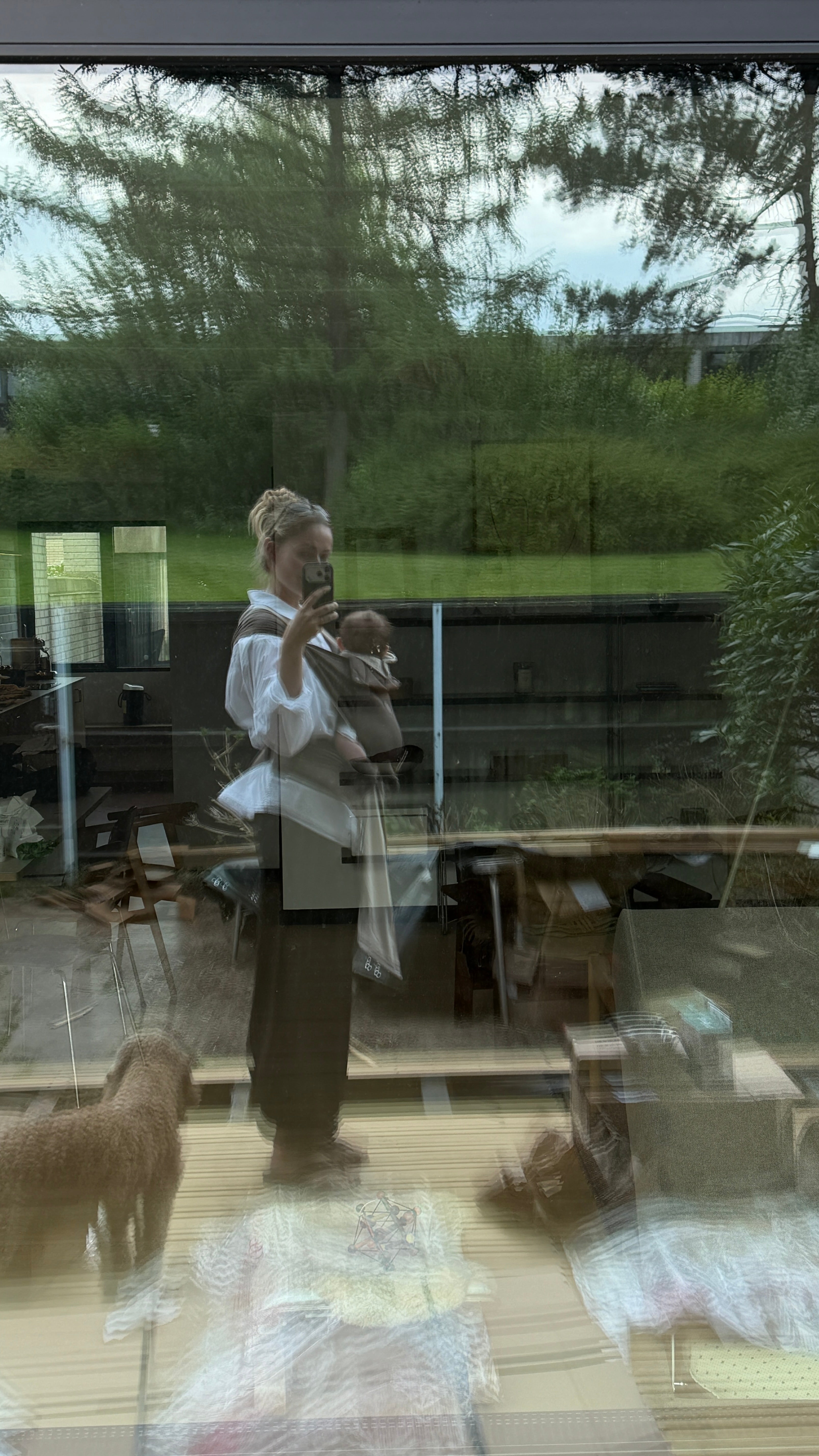 A woman wearing a white blouse and dark skirt stands inside a glass-front room, holding a baby carrier with a baby inside. Large windows behind her show a green outdoor garden, and there are reflections and plastic sheeting on the glass.