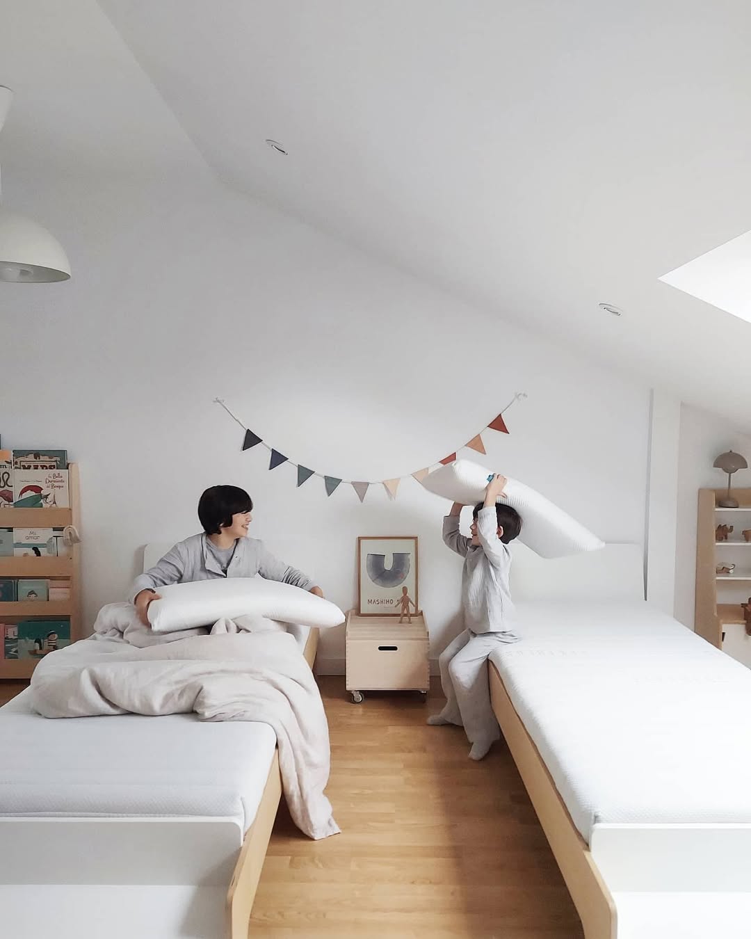 Two people playing with pillows in a modern bedroom with wooden floors and white walls.
