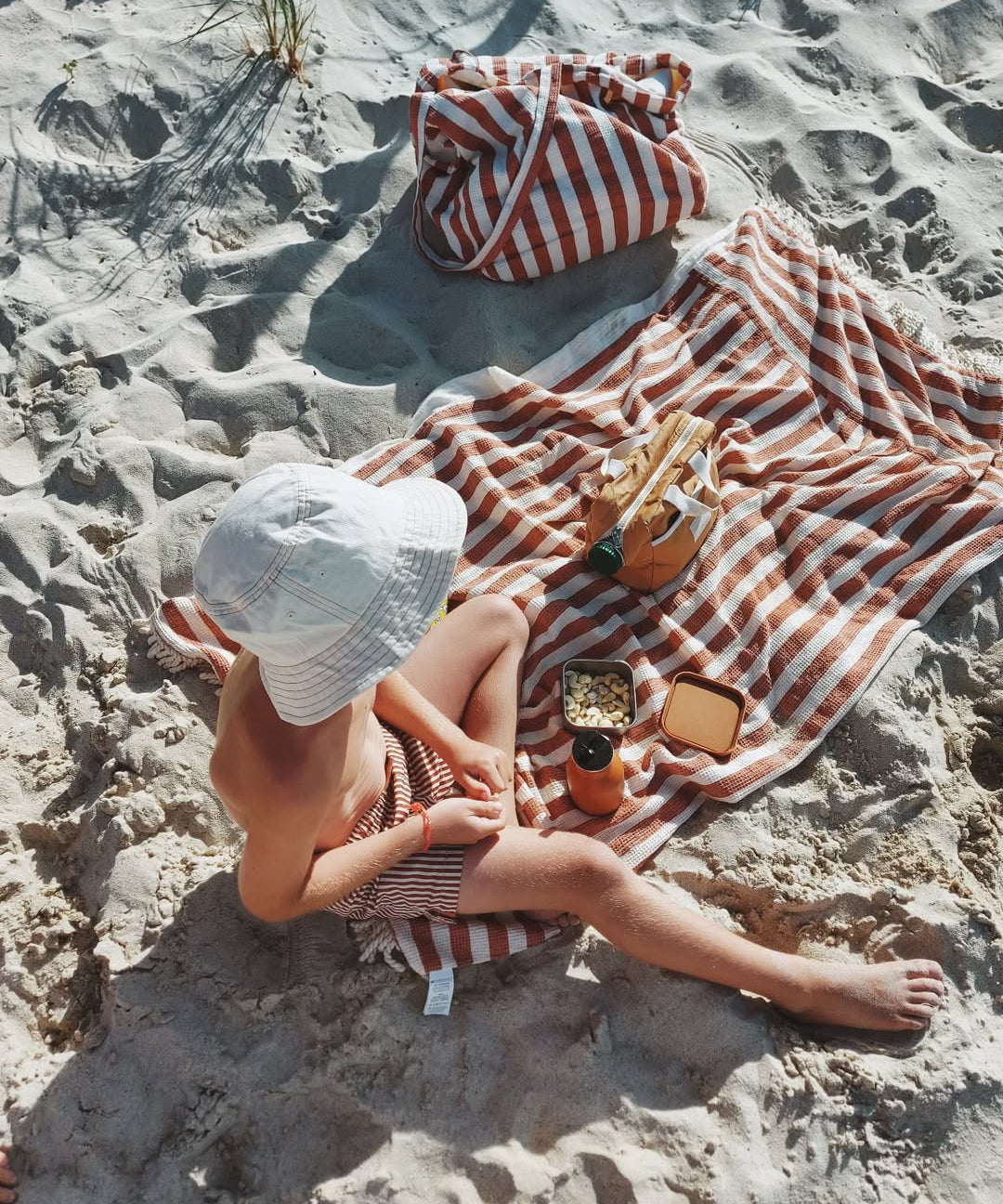 Person sitting on a striped towel on a sandy beach with a hat and sunglasses.