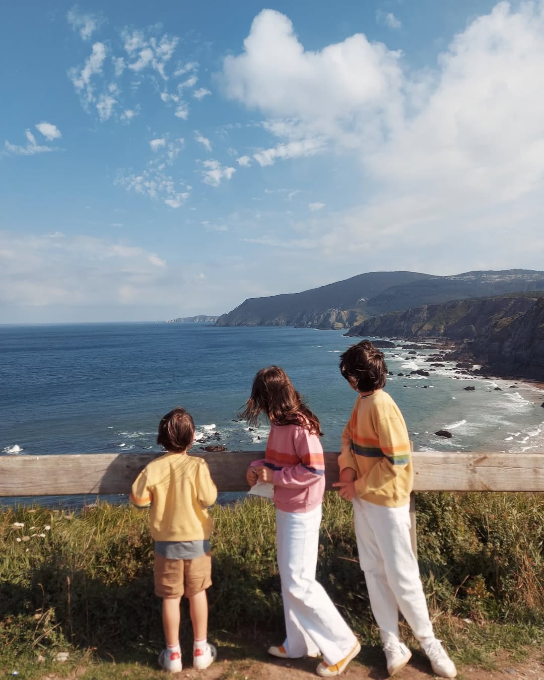 Three children standing on a cliff overlooking a coastal landscape with mountains and a clear sky.