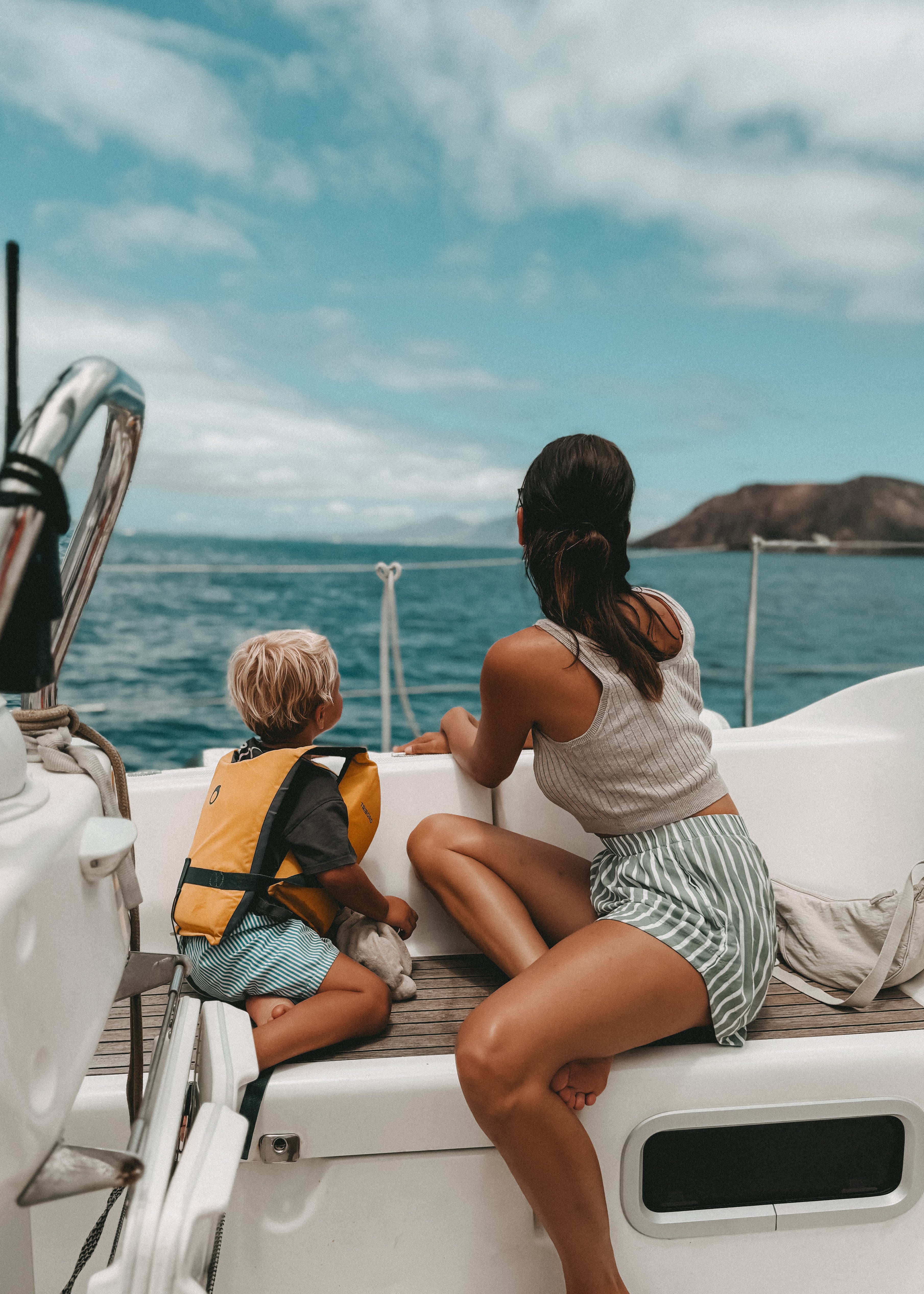 A woman and a child sitting on the deck of a white sailboat, viewed from behind as they look out toward the open ocean under a blue sky.