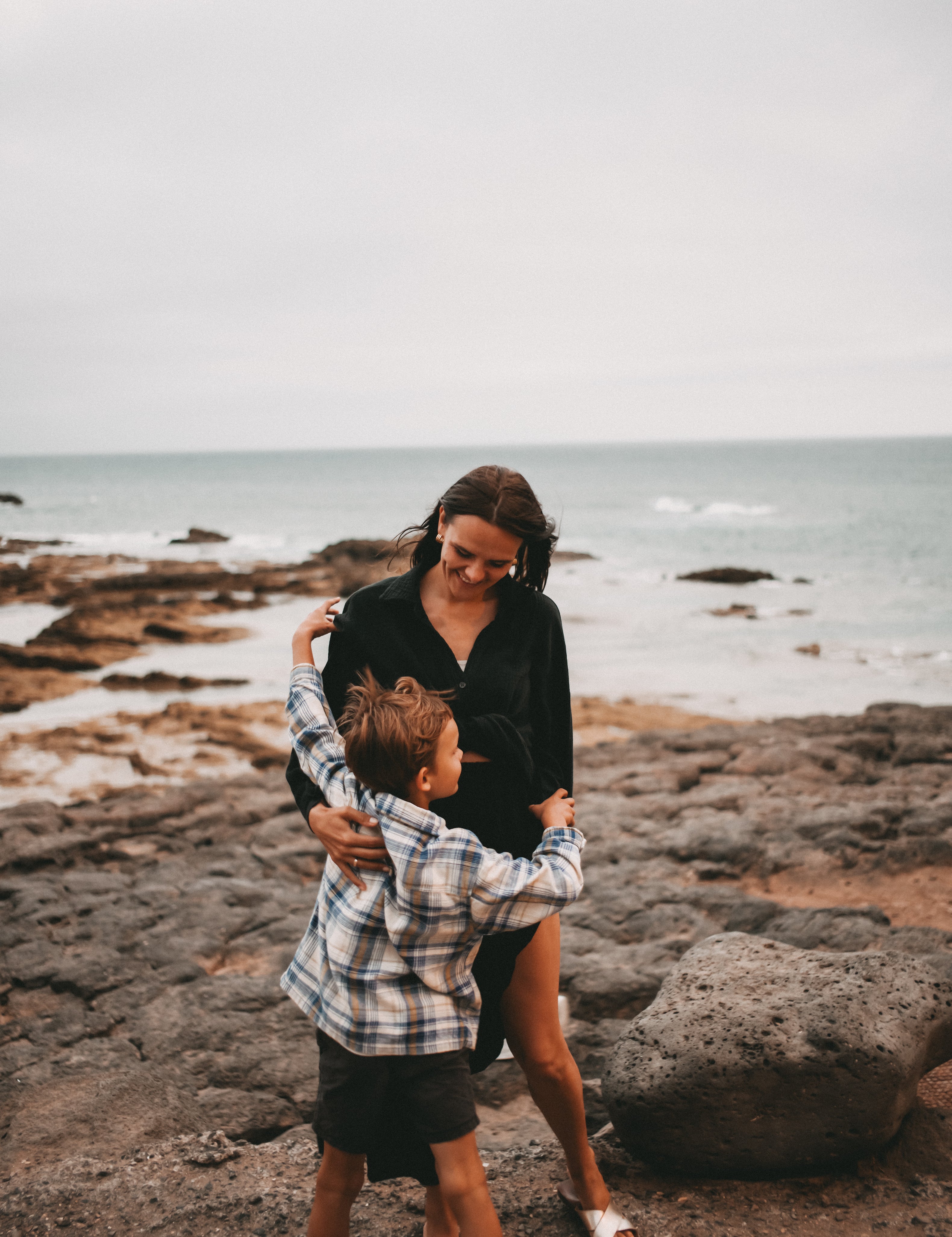 A woman in a black shirt and a young boy in a plaid shirt hugging on a rocky beach with the ocean in the background.