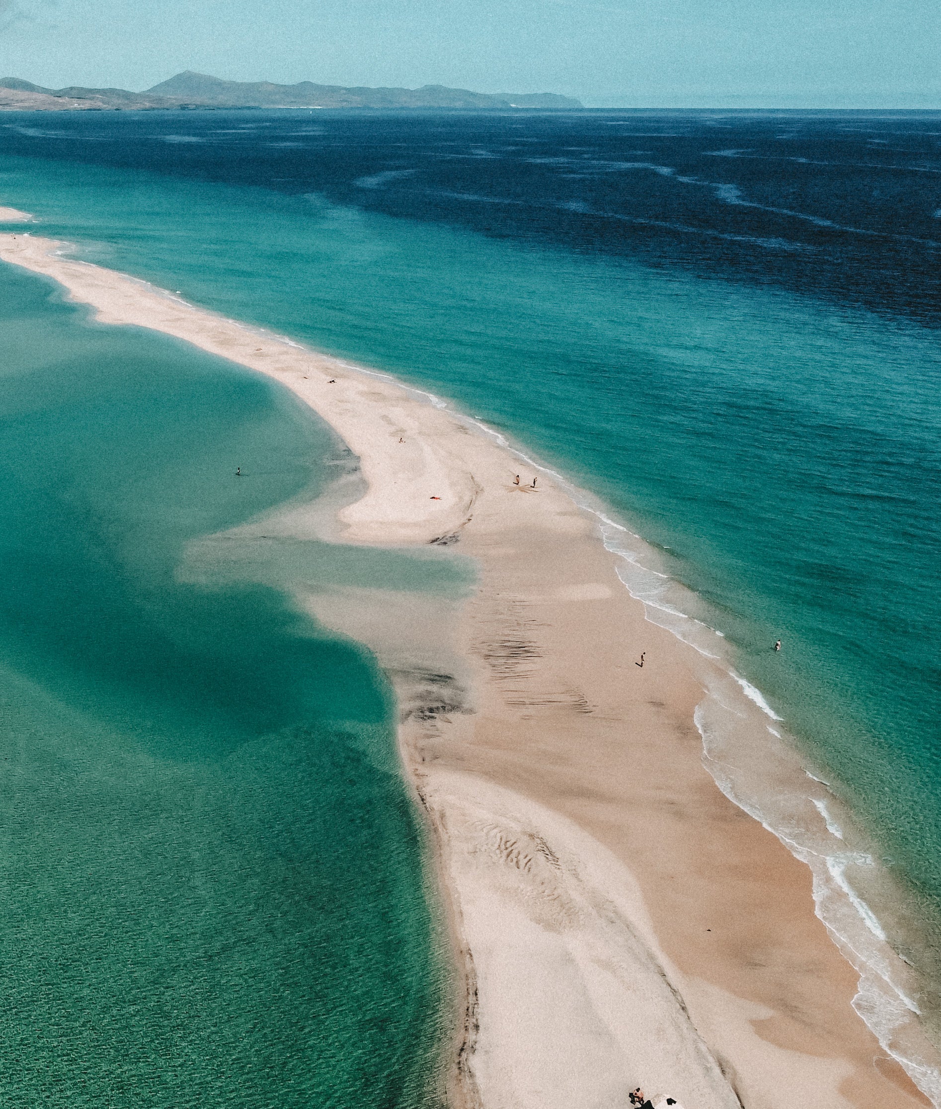 Aerial view of a long sandy spit or sandbar winding between deep blue ocean waters under a pale blue sky, with a few tiny people visible along the shoreline.