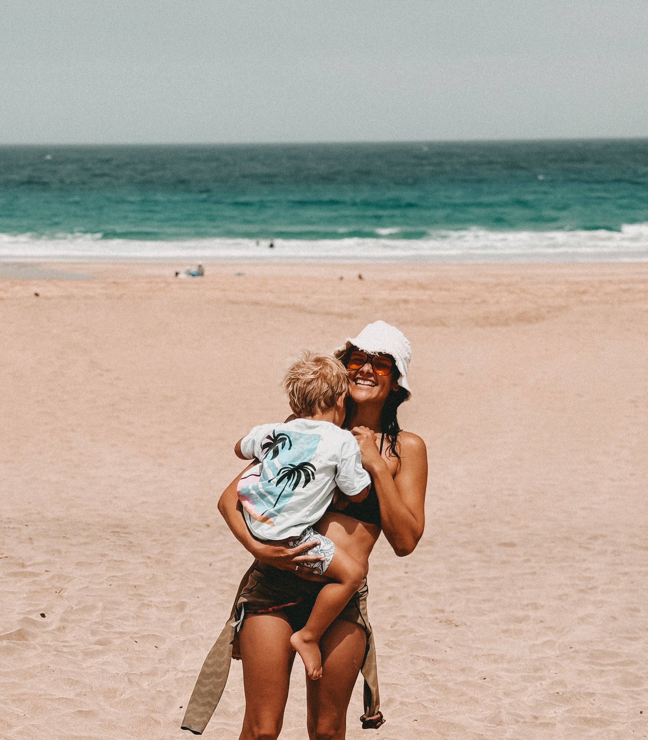 A woman wearing a white bucket hat and sunglasses carries a young child on her shoulders while standing on a sandy beach with the ocean in the background.