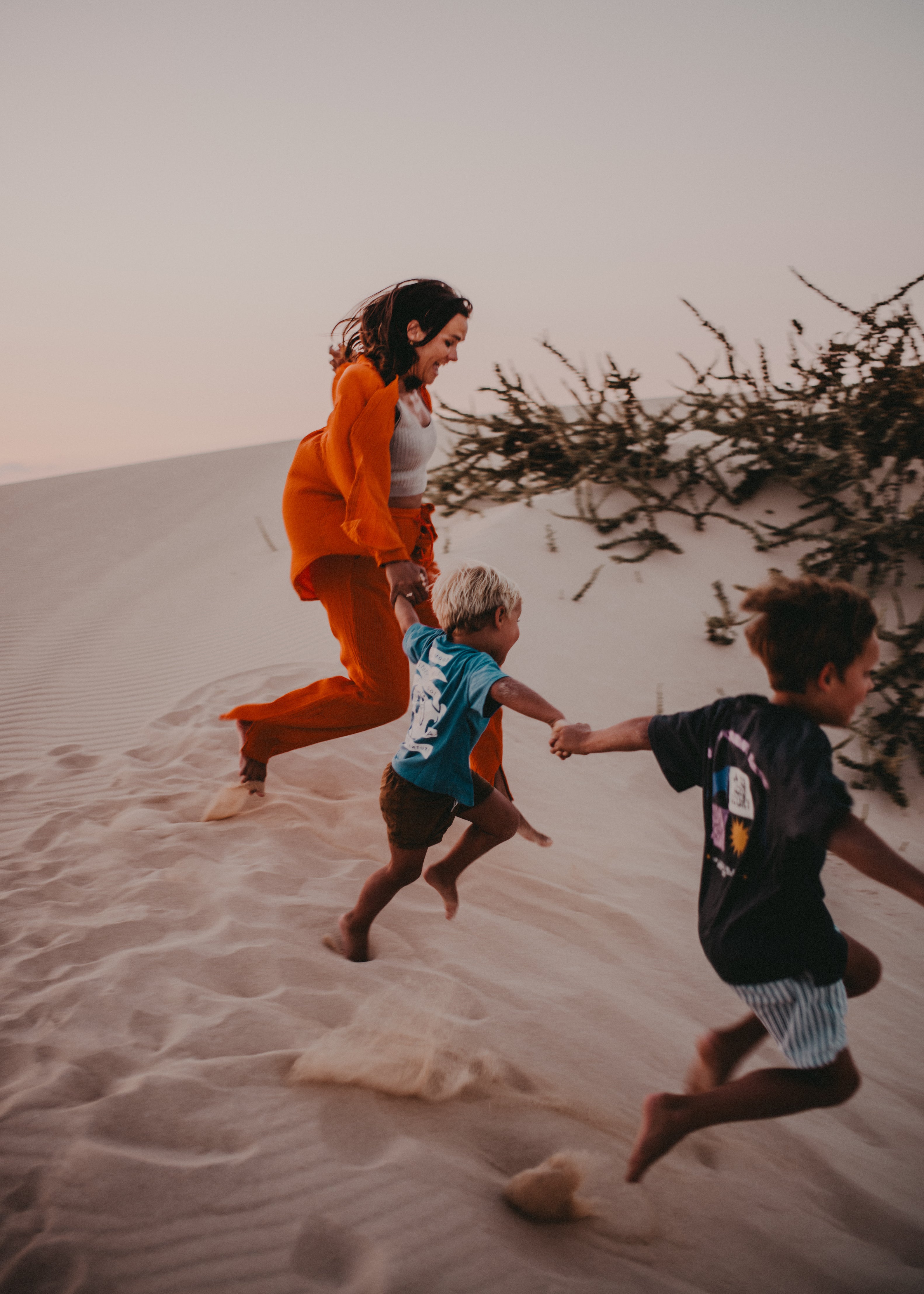A woman in a bright orange tracksuit runs hand-in-hand with two young children across a sandy dune during golden hour, with sparse desert shrubs in the background.