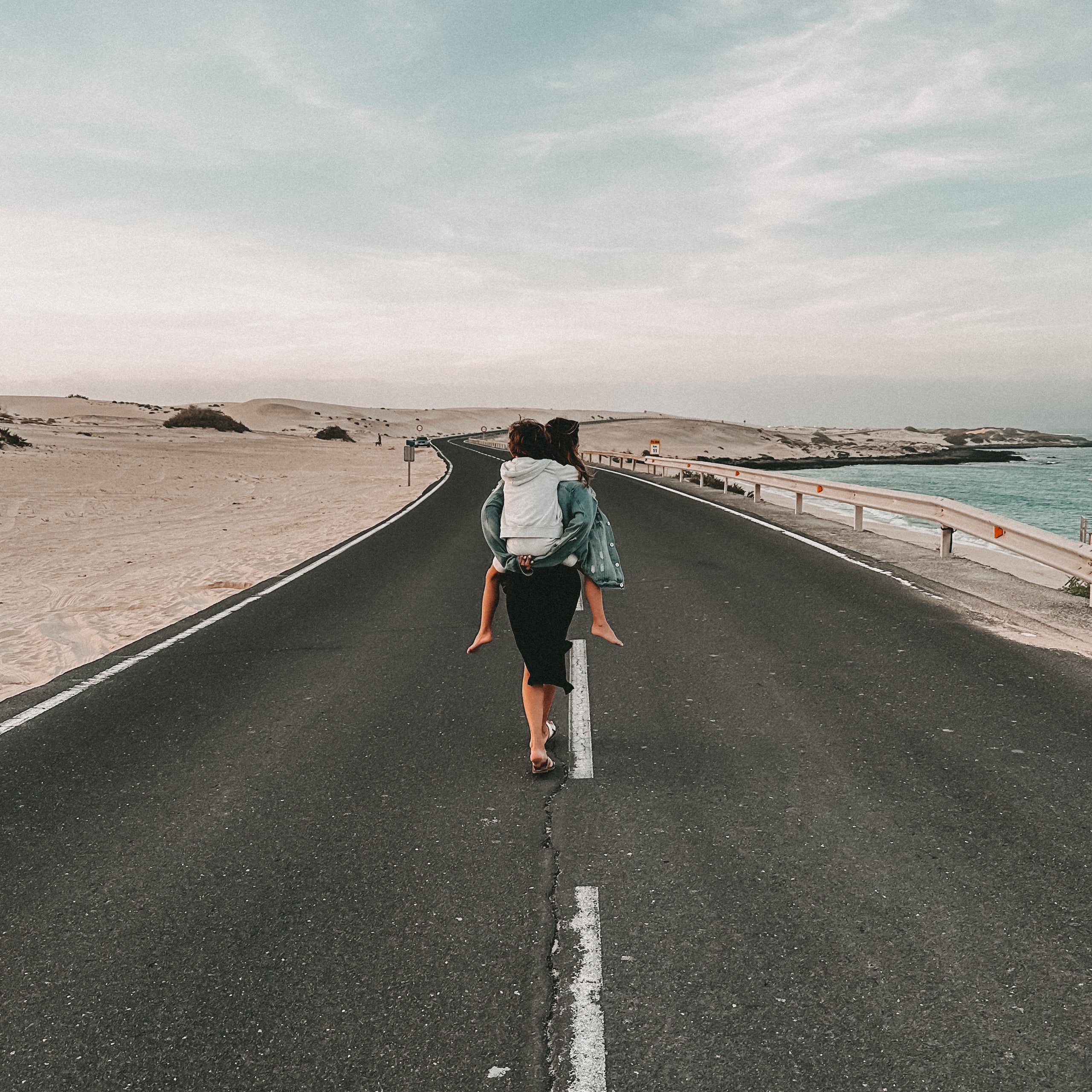 A person with a white backpack walks away on a paved road between sandy terrain and the sea under a pale blue sky.