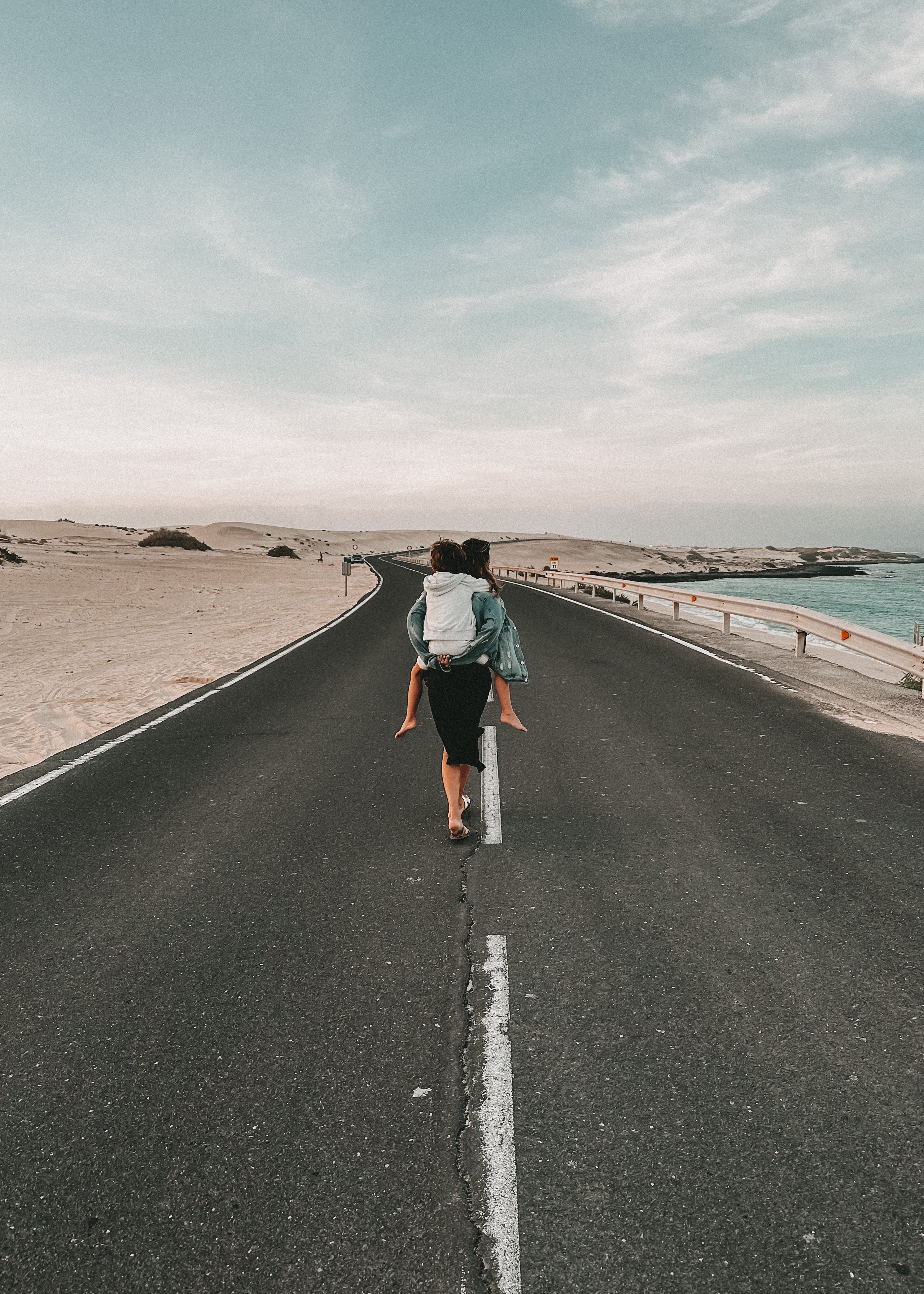 A person with a white backpack walks away on a paved road between sandy terrain and the sea under a pale blue sky.