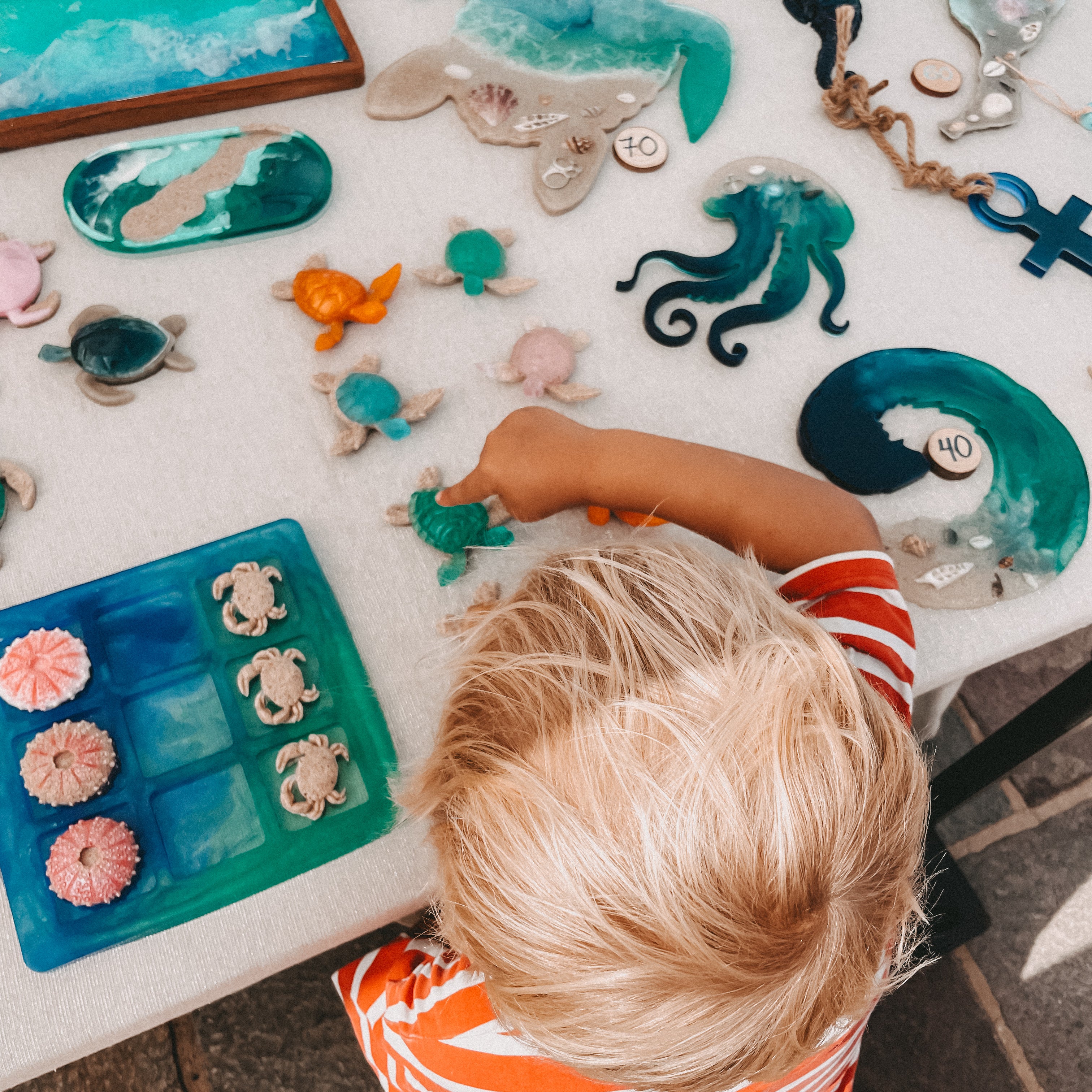 A child in an orange and white striped shirt sits at a table surrounded by handmade sea-themed resin crafts, including sea turtles, octopus figures, shells, and a blue tray, viewed from above.