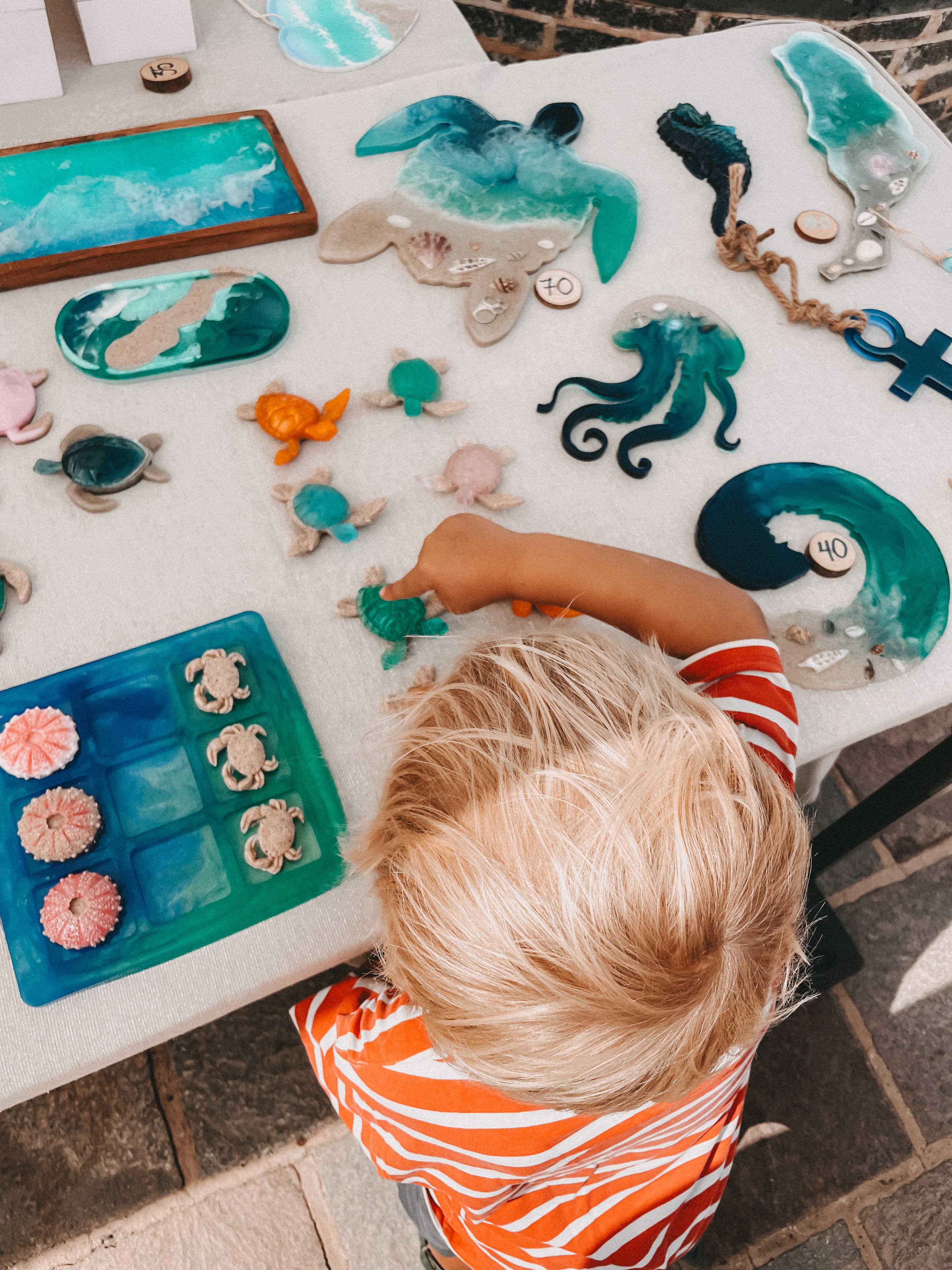 A child in an orange and white striped shirt sits at a table surrounded by handmade sea-themed resin crafts, including sea turtles, octopus figures, shells, and a blue tray, viewed from above.