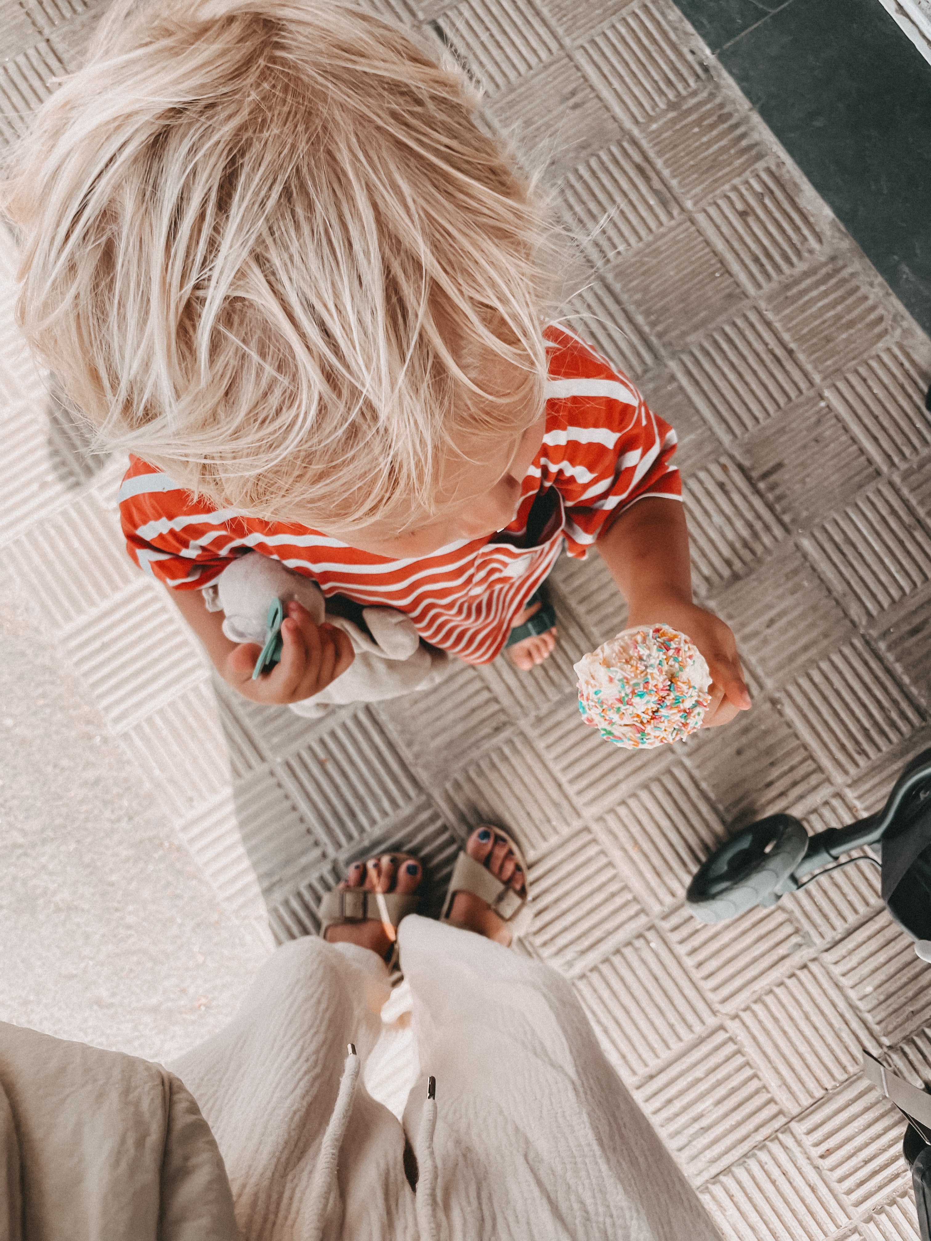 A top-down view of a young child with blonde hair wearing a red and white striped shirt, holding a sprinkle-covered ice cream cone on a textured beige tiled floor.