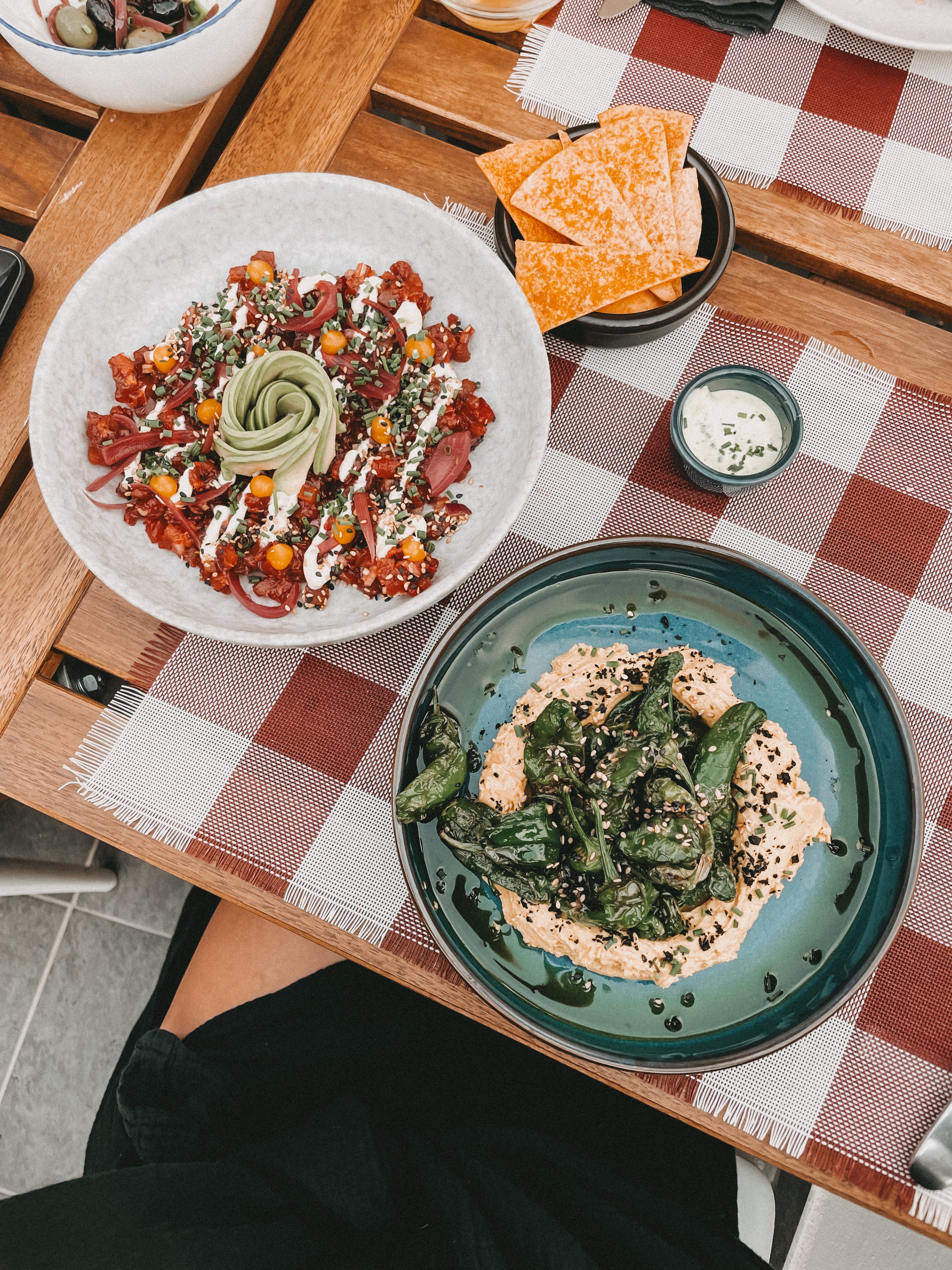 Two plated dishes on a wooden table with a red-and-white checkered placemat: a white bowl filled with a colorful salad and a teal plate with roasted green vegetables on a beige base, alongside orange chips and a small dipping sauce.