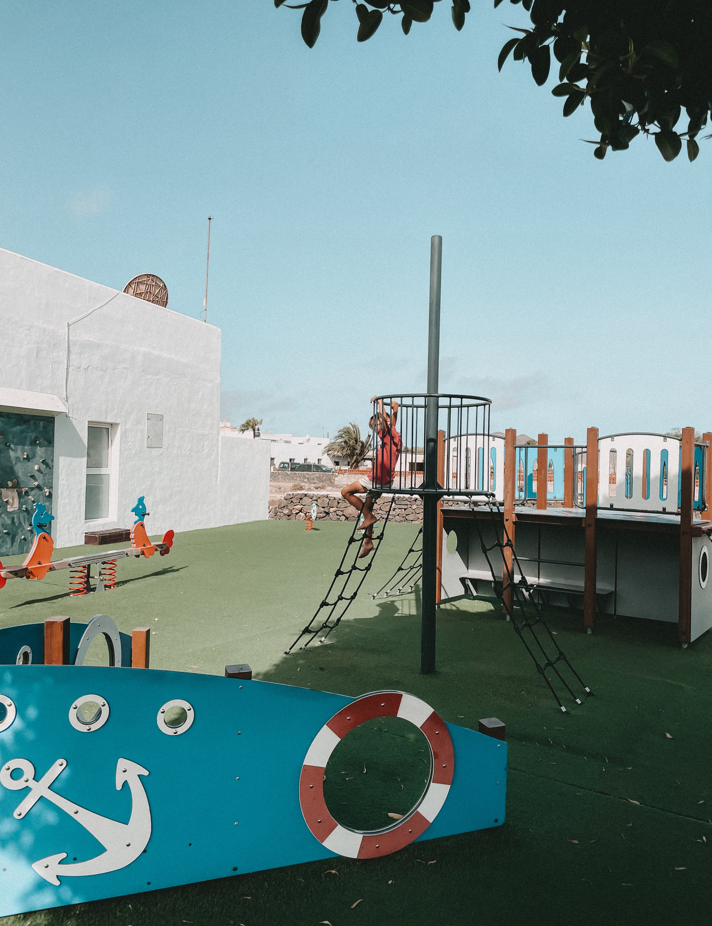 Outdoor playground scene with a child climbing a black ladder structure near a white building, under a blue sky. Playground equipment includes a blue boat-shaped panel with a white anchor and red-and-white life preserver motif, green turf flooring, and wooden railings.