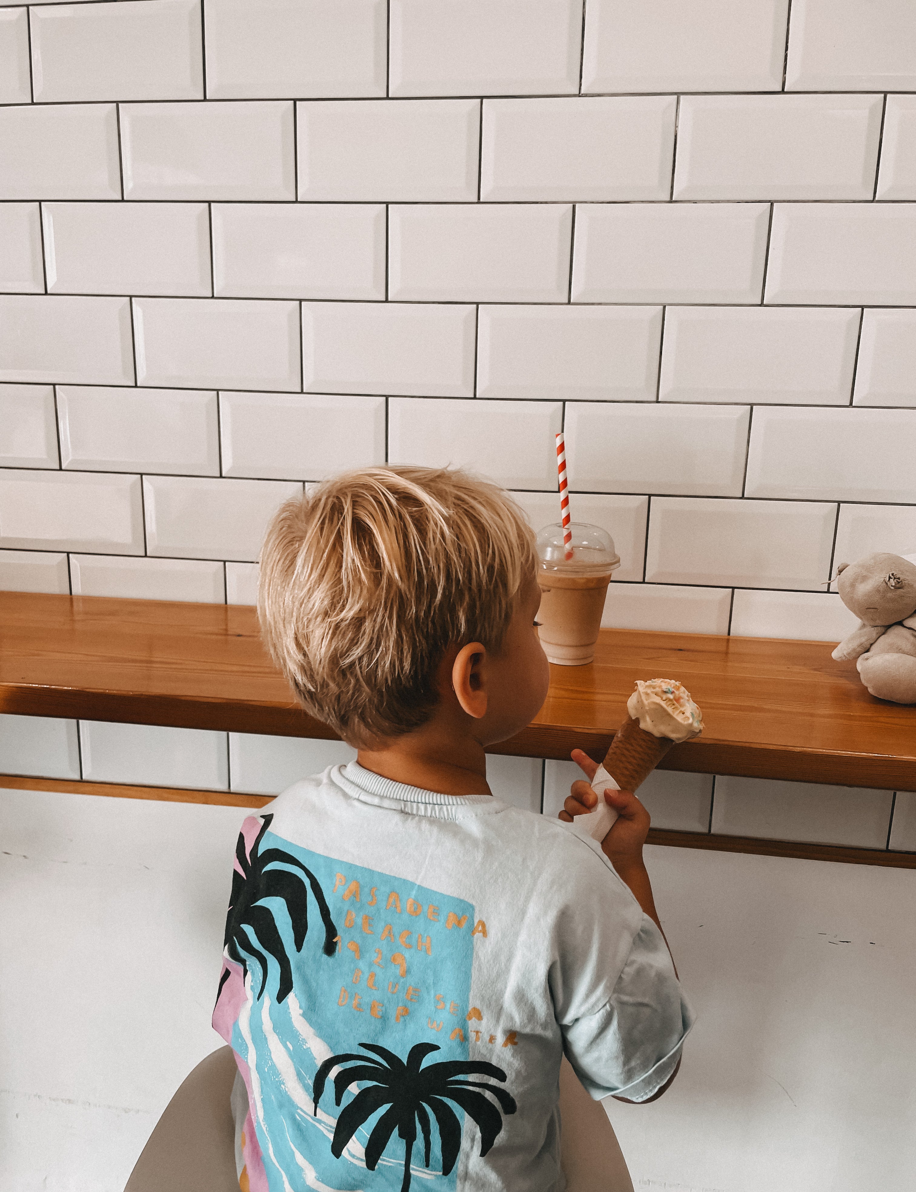 A young child with blonde hair is seen from behind, wearing a light blue t-shirt with a black palm tree graphic and gold text. The child is holding an ice cream cone in a shop setting with a white subway tile wall, a wooden counter, and an iced coffee in the background.
