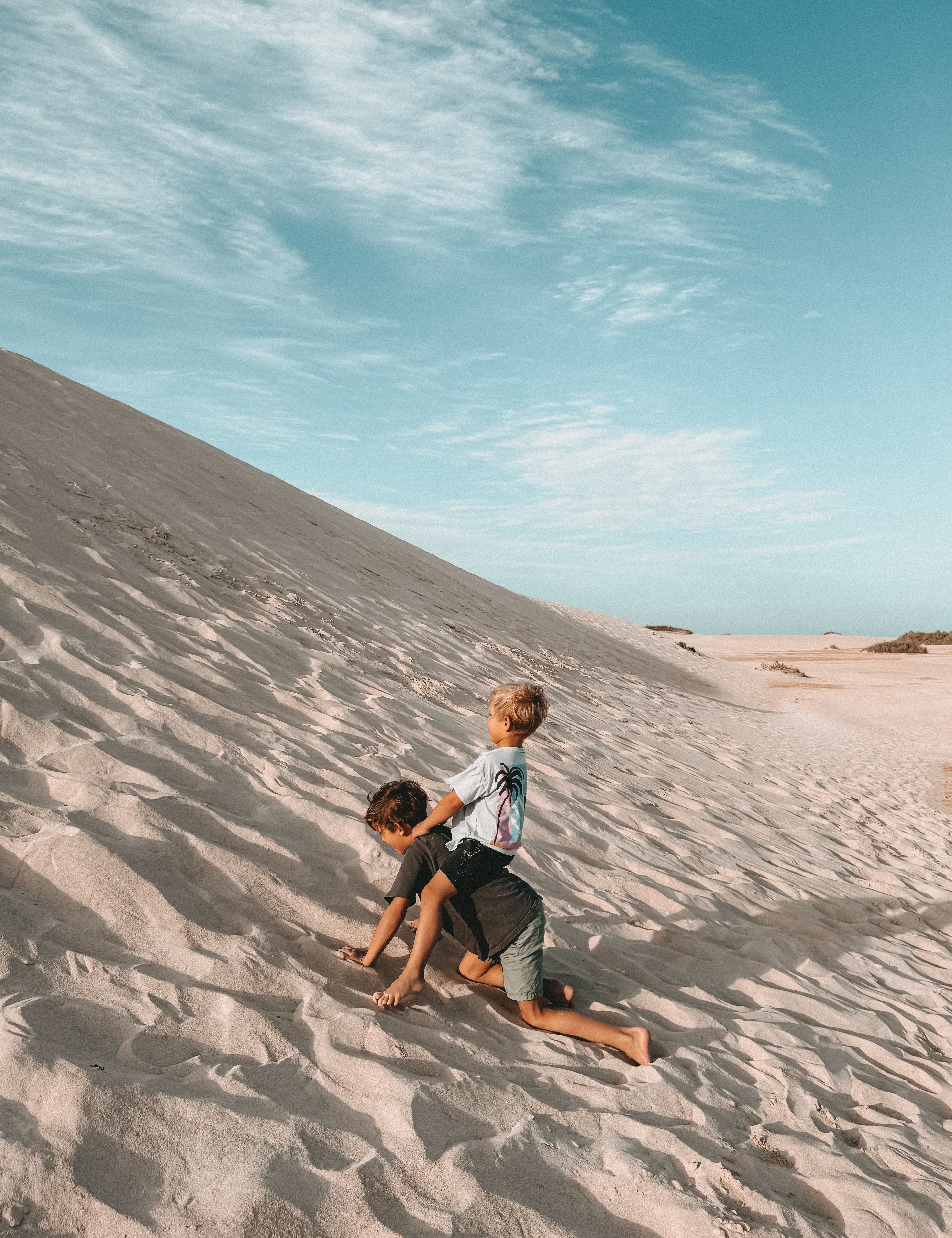 Two children playing on a sandy dune under a bright blue sky with wispy clouds. One child is sitting on the sand while the other stands on their shoulders.